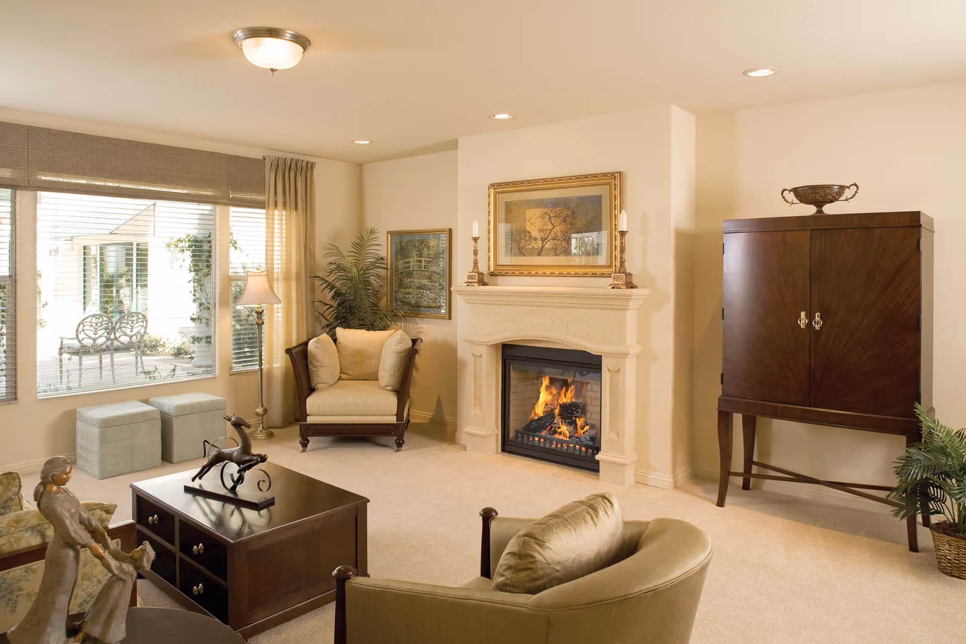 Bright living room with a lit fireplace, armchairs, coffee table, and a large window overlooking a patio.
