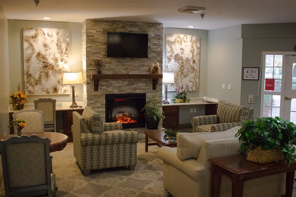A cozy living room area in Woodbury Wellness Center featuring a stone fireplace with an electric fire, a mounted flat-screen TV above it, two large abstract paintings on the wall, two table lamps on wooden desks, several cushioned armchairs and a sofa arranged around a wooden coffee table, and a side table with a green potted plant. There is a door with glass panels on the right side letting in natural light.