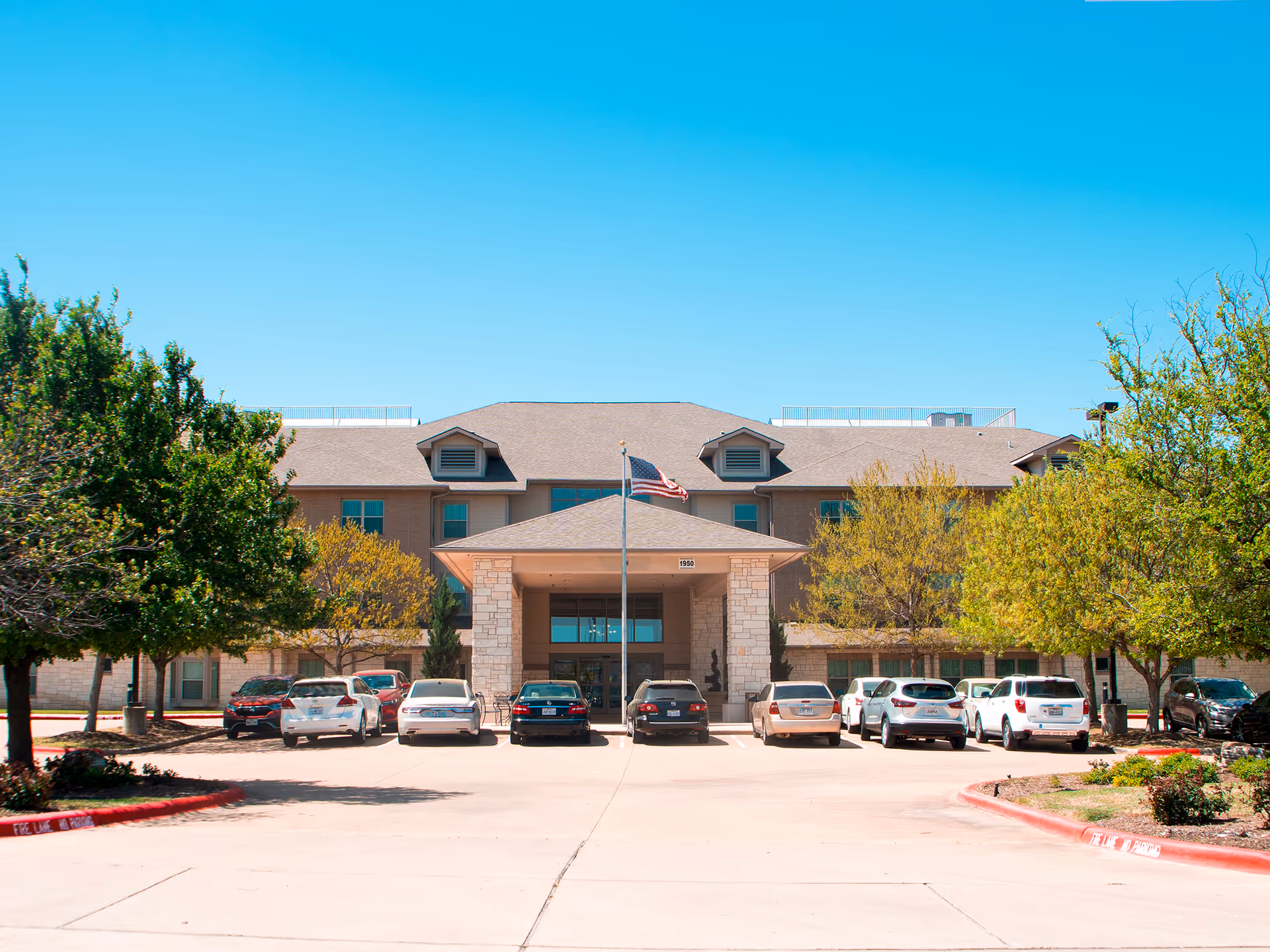 Front entrance of a multi-story retirement facility with parked cars, an American flag, and trees.