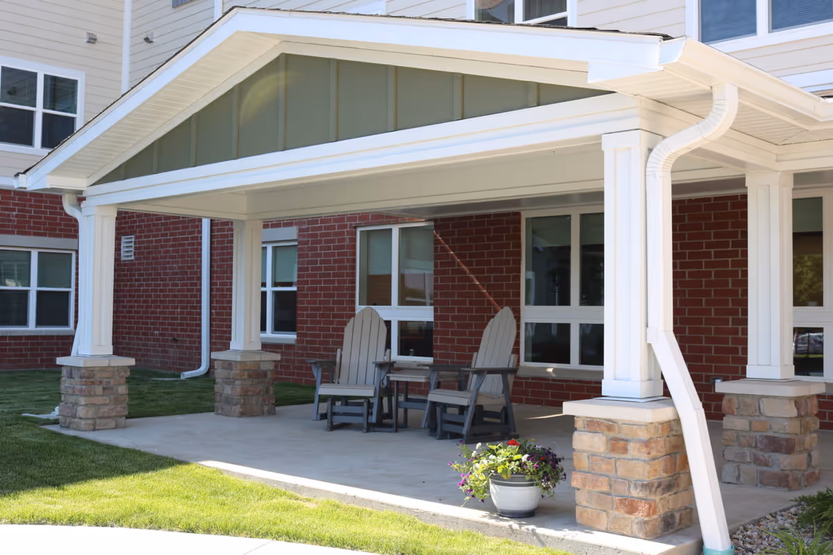 Covered outdoor patio area with two wooden Adirondack chairs and a small table between them, situated in front of a brick building with white-framed windows. A potted plant with colorful flowers is placed on the concrete floor near the edge of the patio.