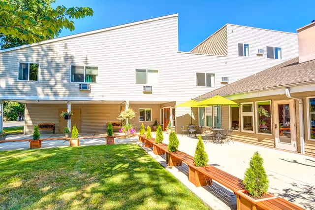 Outdoor patio area of a retirement and assisted living facility with beige siding buildings, green grass, wooden benches with small potted evergreen trees, and a table with a bright green umbrella.