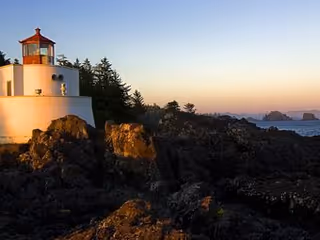 A white lighthouse on a rocky shoreline with trees and distant islands at sunset.
