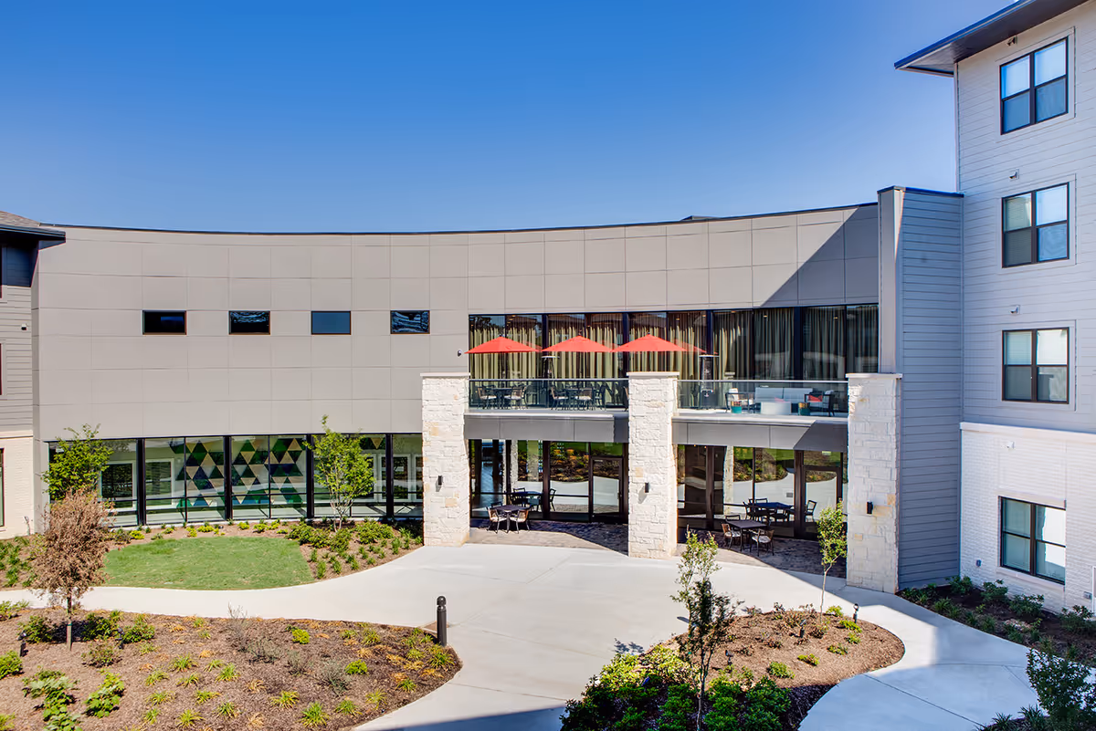 Exterior view of Fountainwood at Lake Houston showing a modern building with large windows, a balcony with red umbrellas, outdoor seating areas, landscaped garden beds, and a clear blue sky.