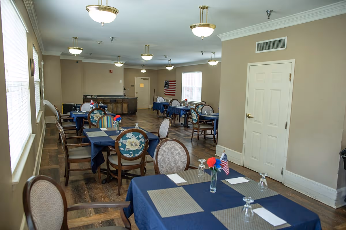 Interior view of a dining room with multiple tables covered in blue tablecloths and set with placemats, napkins, upside-down glasses, and small vases holding red and blue flowers along with small American flags. The room has beige walls, wooden flooring, several windows with blinds, and ceiling lights. There is a door on the right wall and an American flag hanging on the far wall.