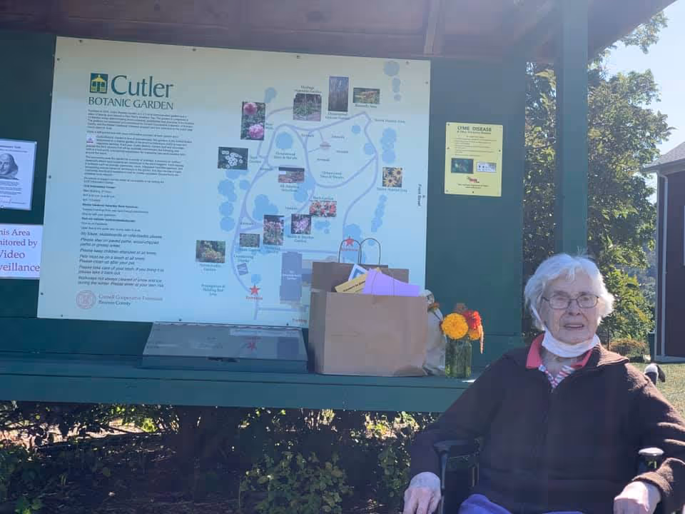 An elderly woman sitting in a wheelchair outdoors near a large informational signboard for Cutler Botanic Garden. The sign includes a map and text about the garden. There are paper bags and a vase with flowers placed on the bench beneath the sign. Trees and a building are visible in the background.