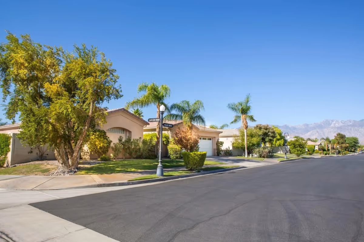 A sunny suburban street with single-story houses, palm trees, and well-maintained lawns under a clear blue sky with mountains in the background.