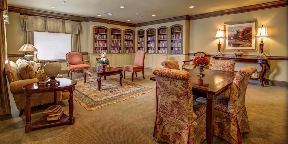 A cozy and elegant living room area with a beige carpet and warm lighting. The room features a sofa, several upholstered chairs, a wooden coffee table, and a dining table with six patterned chairs. There are built-in bookshelves filled with books along the back wall, a window with curtains, table lamps, and a framed landscape painting on the wall.