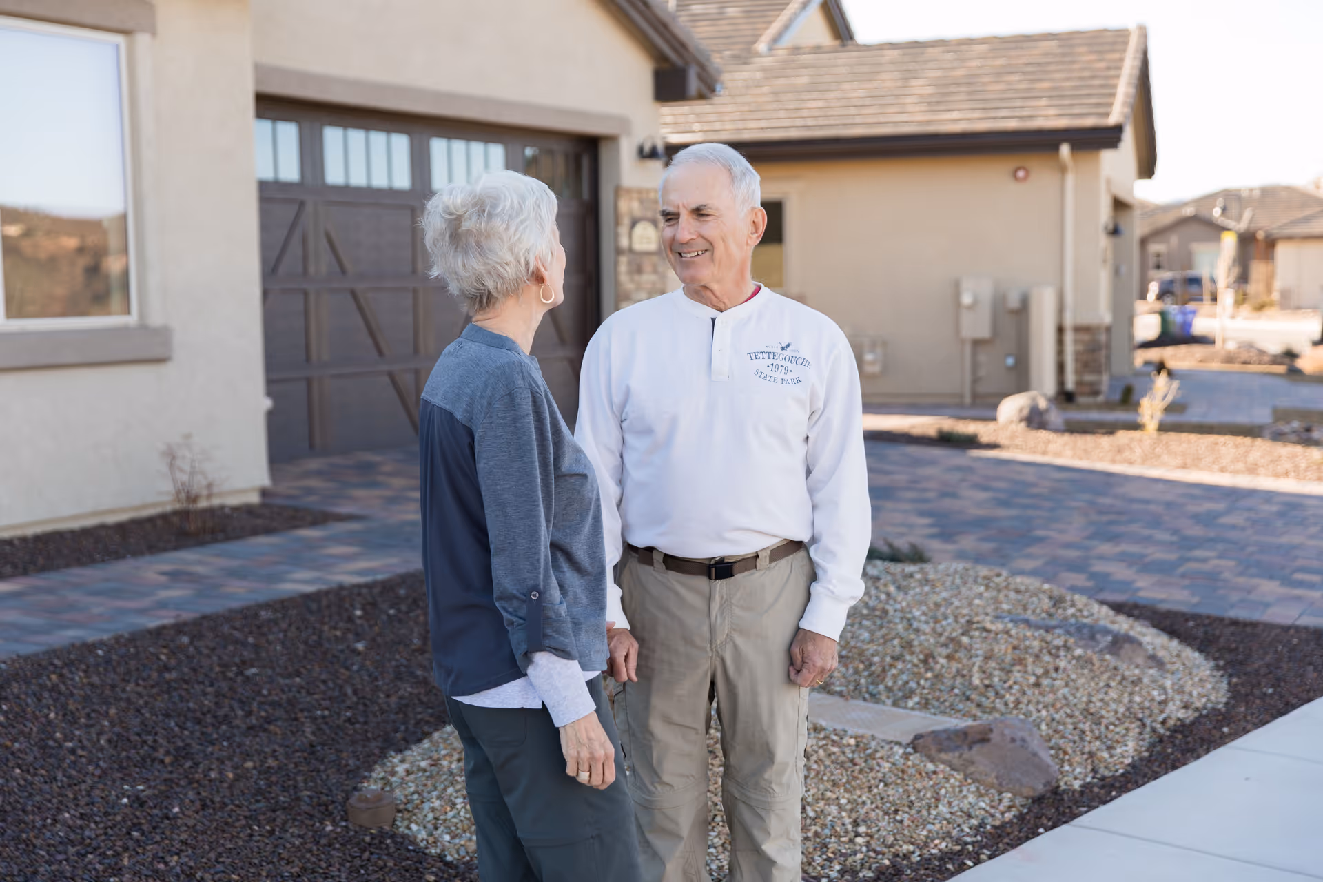 An elderly man and woman standing outside in front of a residential building, engaged in conversation. The setting includes a paved driveway, gravel landscaping, and beige-colored houses with brown roofs in the background.