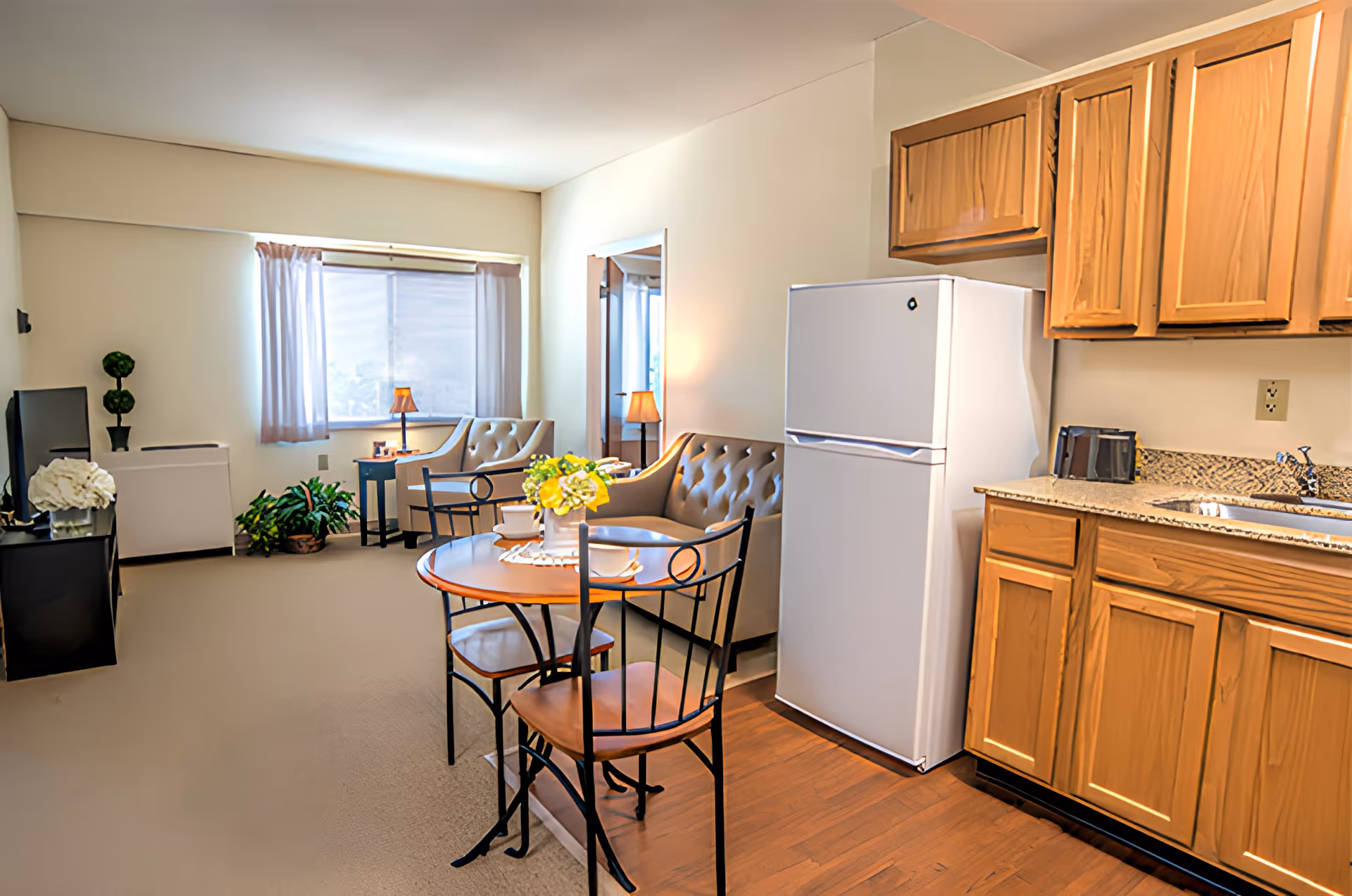 Open-plan studio living area with a small dining table, sofa, TV, and a kitchenette with refrigerator and wooden cabinets.