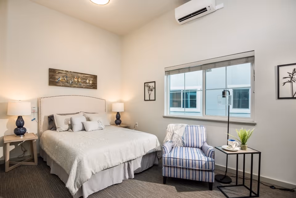 A cozy bedroom with a large bed featuring a beige upholstered headboard and light-colored bedding. There are two bedside tables with matching blue lamps on either side of the bed. A striped armchair with a throw blanket is positioned next to a small black side table holding a cup and a small potted plant. A floor lamp stands beside the chair. The room has a window with blinds, light-colored walls, and a wall-mounted air conditioning unit above the window. Artwork depicting birds and plants decorates the walls.