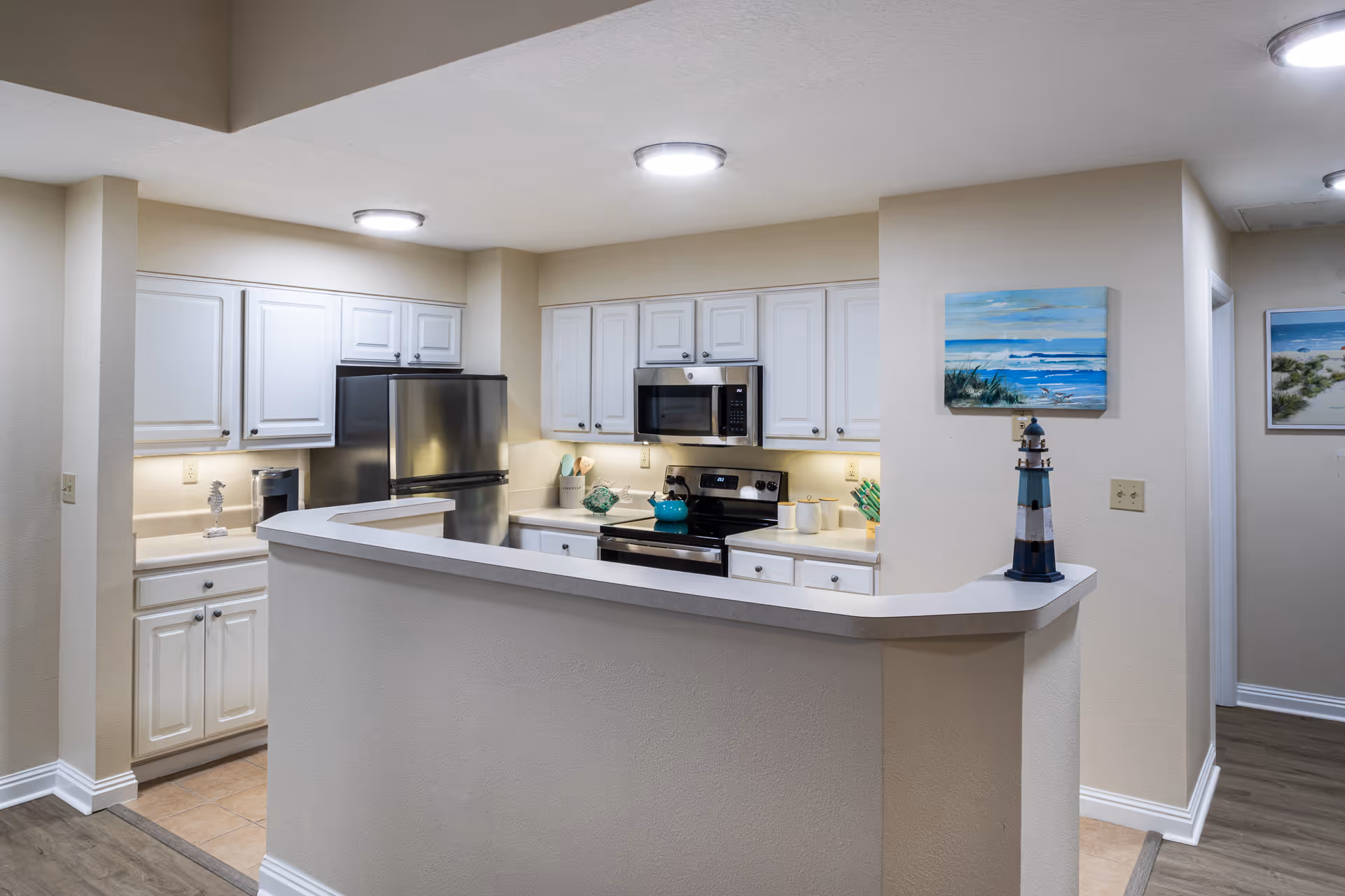 A modern kitchen with white cabinets, stainless steel refrigerator, stove, and microwave. The kitchen has a light beige countertop with a small lighthouse decoration on the corner. The walls are painted light beige, and there are two paintings of beach scenes on the walls. The floor is a combination of tile and wood.