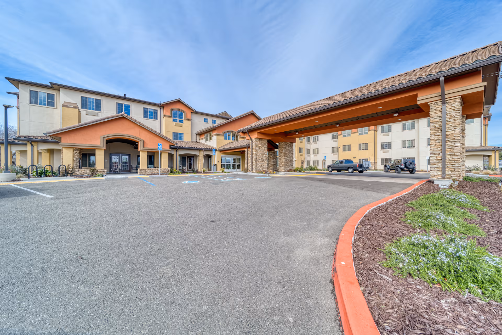 Front entrance of a multi-story senior living building with a covered porte-cochere, parking area, and landscaped curb.