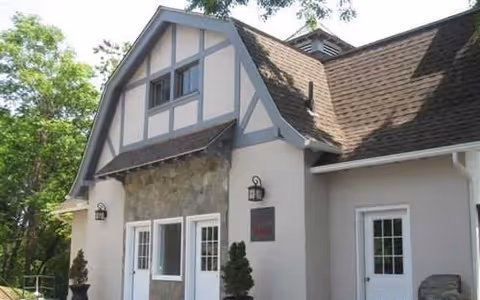 Exterior view of a building with a steep roof, stone and stucco facade, two white doors with glass panes, wall-mounted lantern lights, and some greenery around.