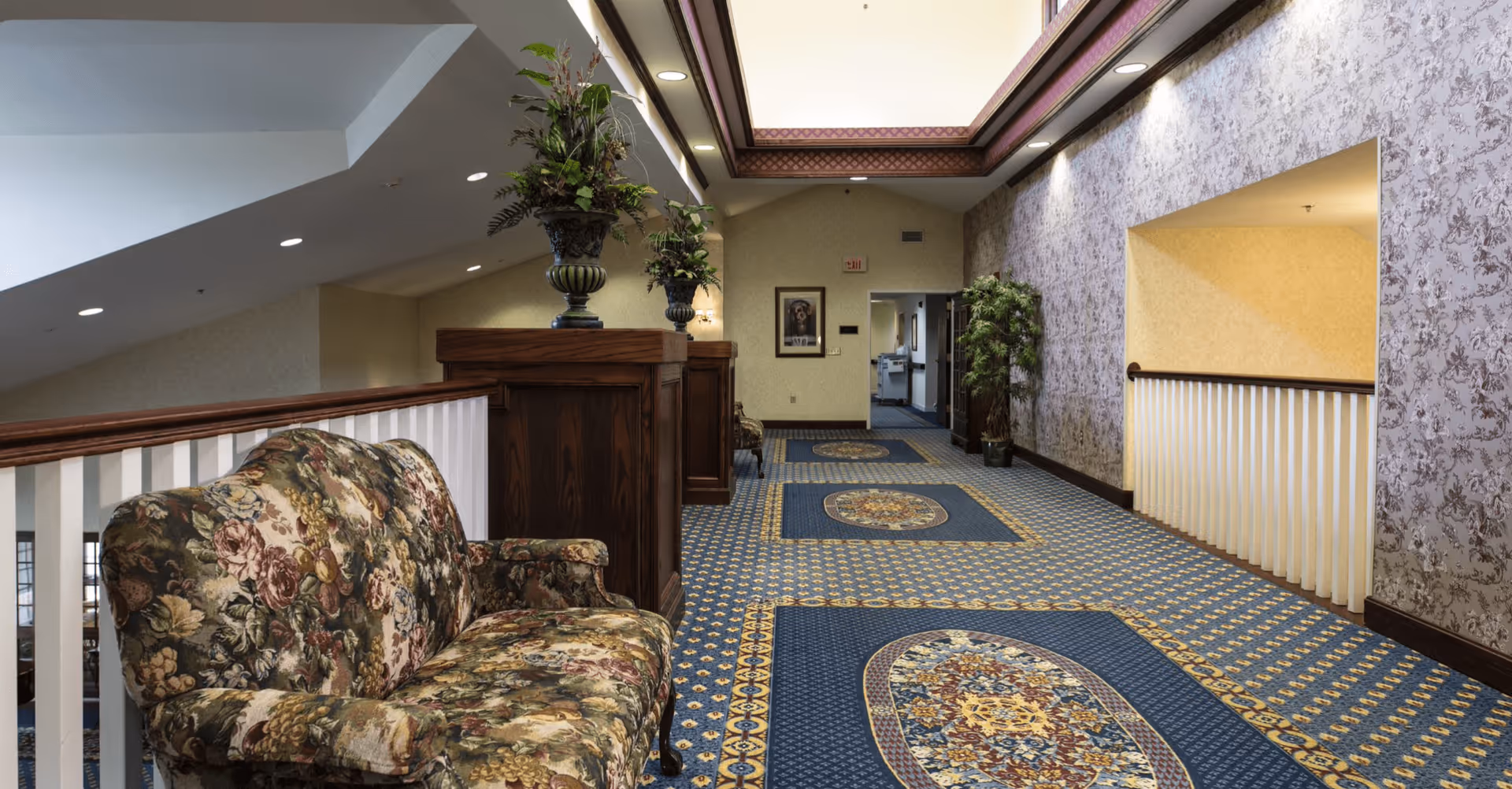 A carpeted hallway in a senior living facility with floral patterned wallpaper and a floral upholstered sofa along the left side. The hallway features decorative wooden railings, potted plants on wooden stands, and recessed lighting in the ceiling.