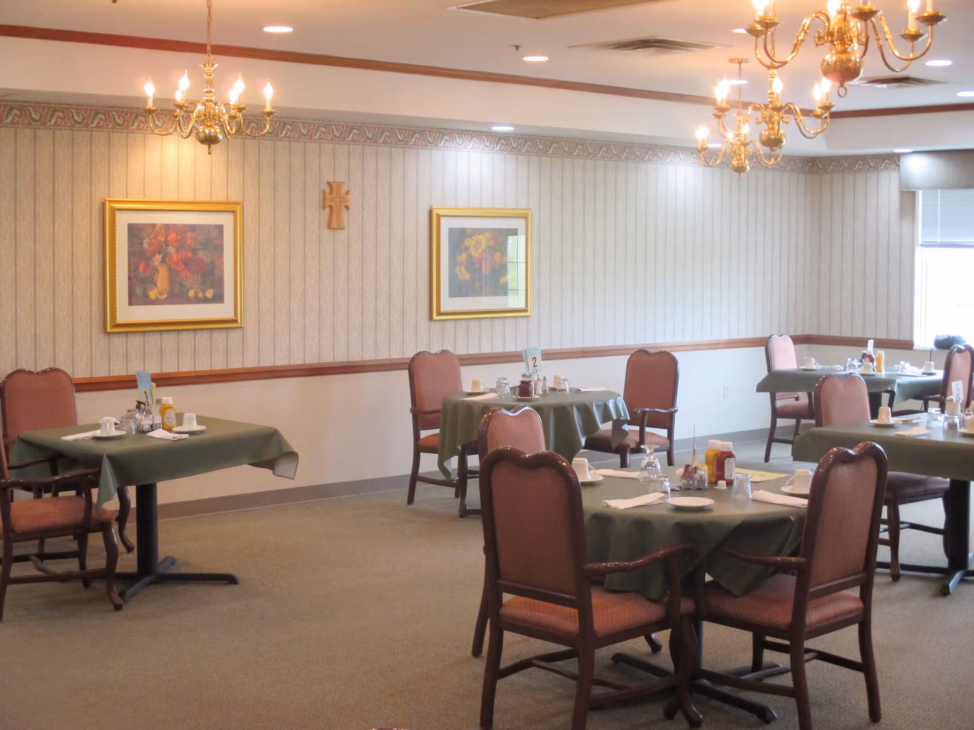 A dining room with several tables covered in green tablecloths, each set with cups, plates, napkins, and condiments. The room has beige wallpaper with a decorative border near the ceiling, two framed floral paintings, and a wooden cross on the wall. There are chandeliers hanging from the ceiling and windows letting in natural light.