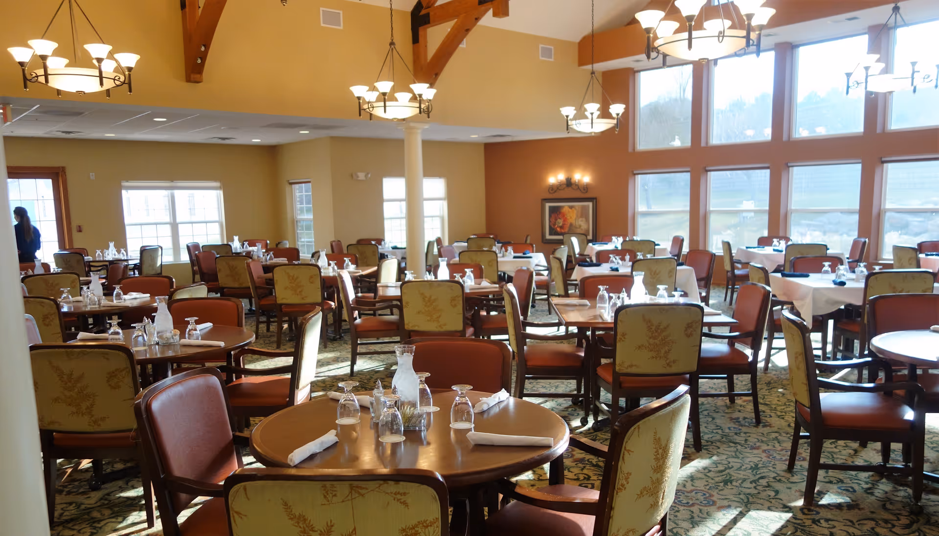 A spacious dining room with multiple round and rectangular tables set with glassware, napkins, and water pitchers. The room features large windows allowing natural light to fill the space, high ceilings with wooden beams, and several chandeliers providing additional lighting. The chairs have cushioned seats and backs with a floral pattern.