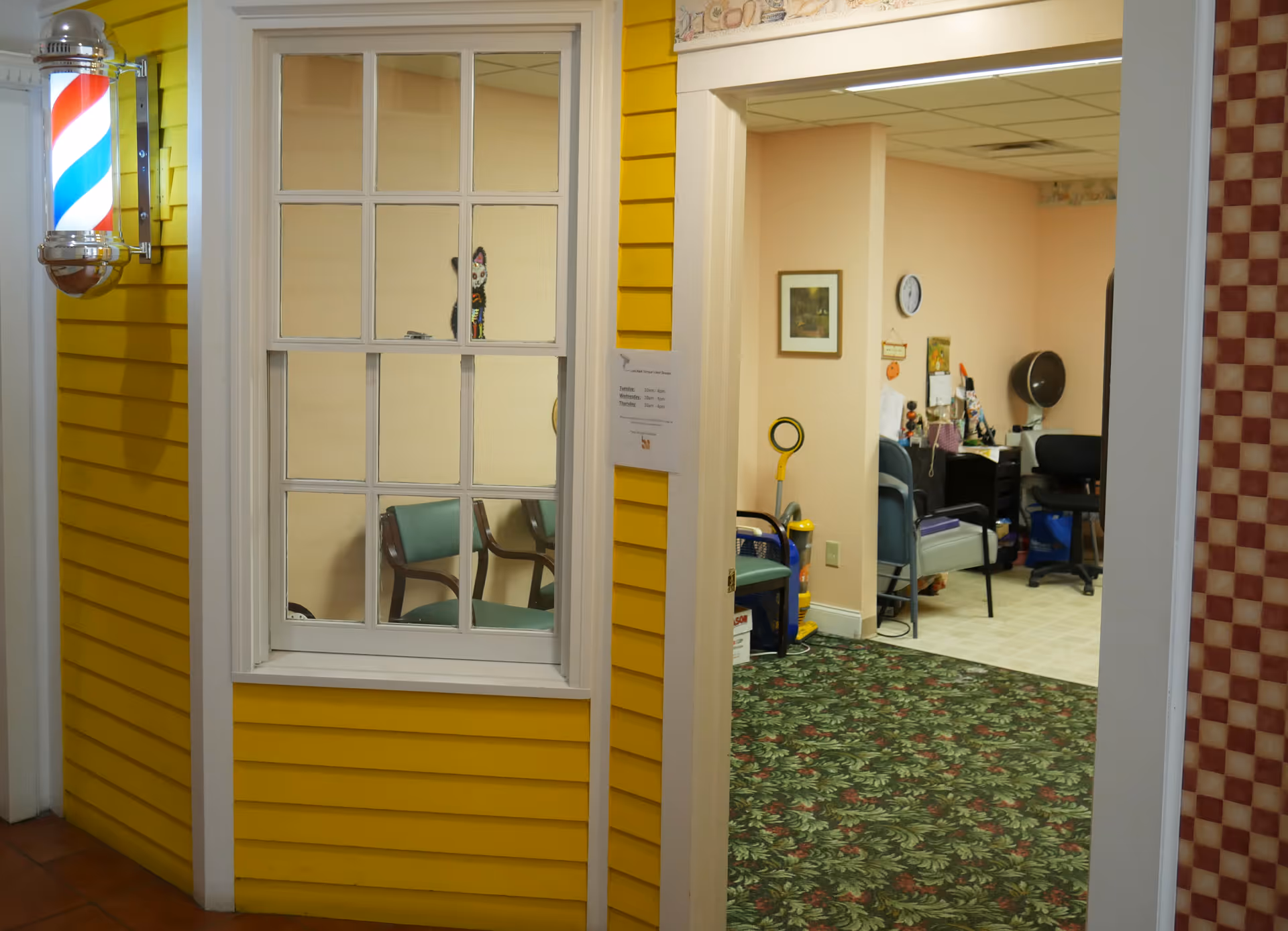 Interior view of a senior living facility showing a yellow wall with a barber pole light fixture next to a window. Through the window and open doorway, a room with chairs, a clock on the wall, and various items including a hair dryer and cleaning supplies is visible.