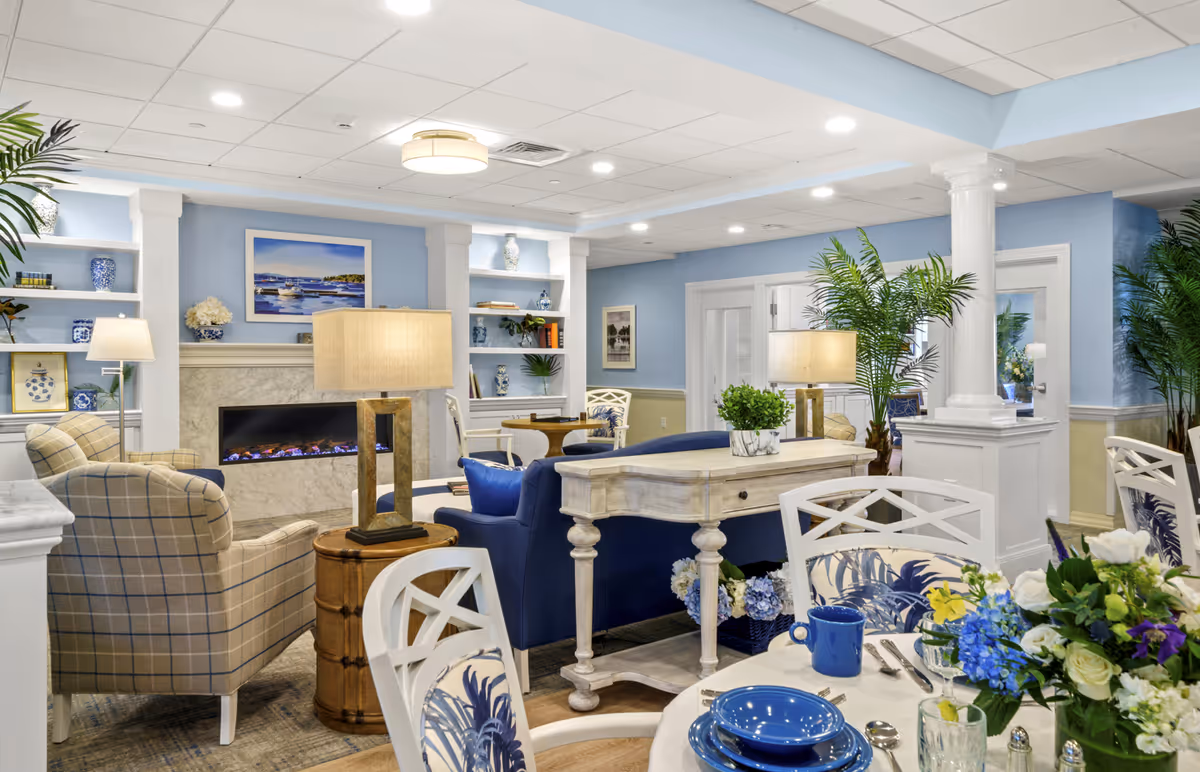 Bright coastal-themed common lounge with sofas, armchairs, a fireplace, a set dining table and decorative plants.