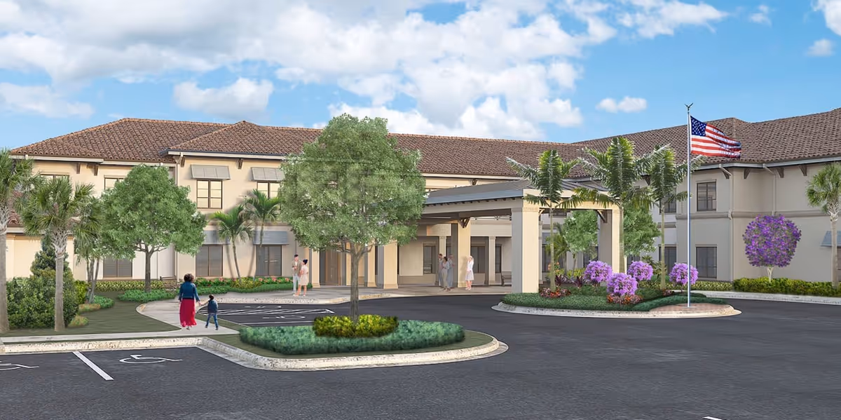 Exterior view of The Capstone at Royal Palm Senior Living facility showing a two-story building with a tiled roof, an entrance canopy, landscaped greenery including trees and purple flowering bushes, an American flag on a flagpole, and a few people walking near the entrance.