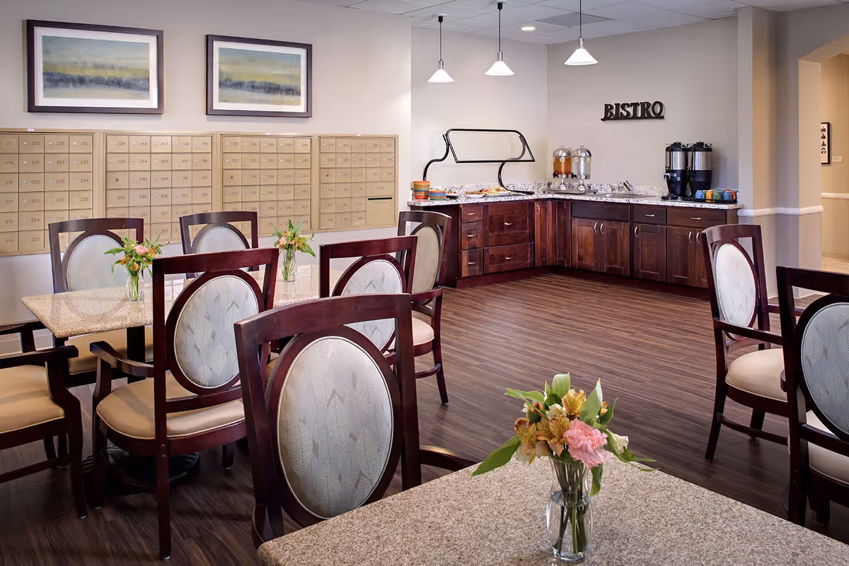 A cozy dining area with several tables and chairs featuring wooden frames and cushioned seats. Each table has a small vase with flowers. In the background, there is a buffet counter with a sign that reads 'BISTRO' on the wall above it. The buffet area has beverage dispensers and plates. On the left wall, there are multiple mailboxes and two framed landscape paintings above them. The floor is wooden, and the lighting is soft with ceiling lights and pendant lamps.