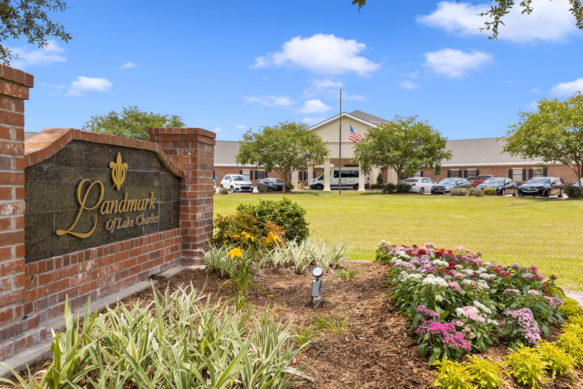 View of the front exterior of Landmark of Lake Charles facility with a brick sign in the foreground surrounded by flowers and greenery, and the building with parked cars and an American flag in the background under a partly cloudy sky.
