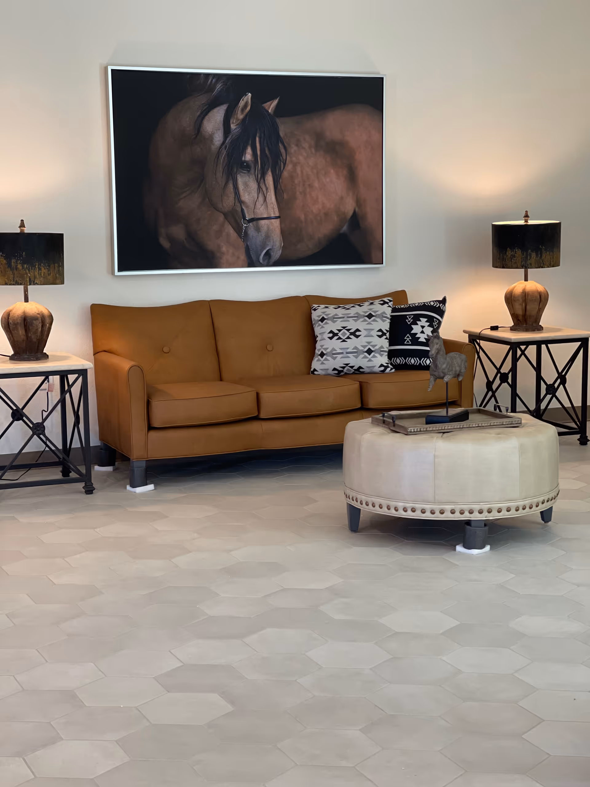 A seating area with a brown leather sofa, patterned pillows, a round ottoman, side tables with lamps, and a large horse painting on the wall.