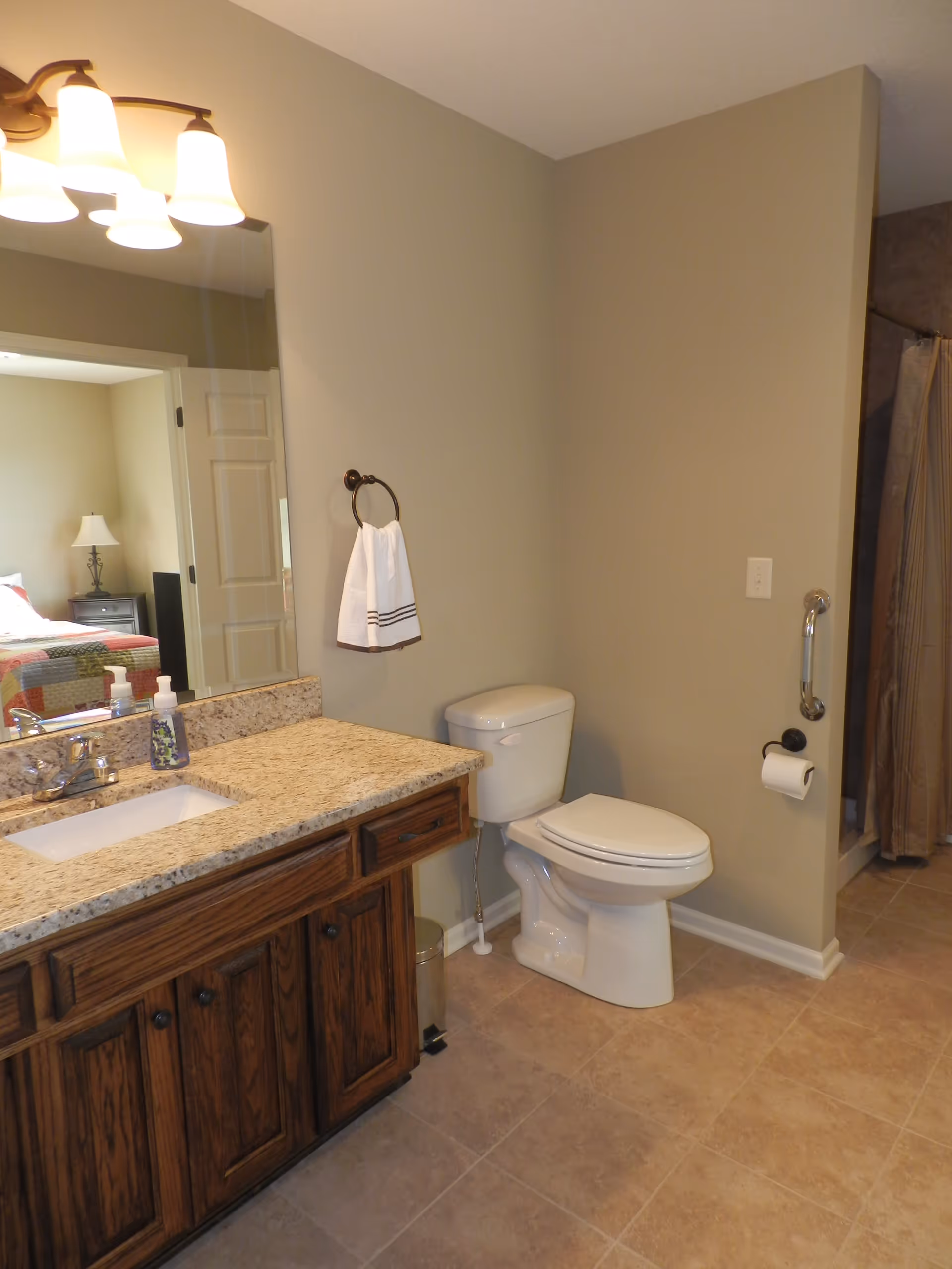 A bathroom with a granite countertop vanity featuring a sink, soap dispenser, and hand soap. There is a large mirror above the vanity with three light fixtures. A white towel hangs on a ring next to the toilet. The toilet is positioned beside a wall with a grab bar and a toilet paper holder. In the background, a shower with a curtain is visible, and a bedroom with a bed and lamp can be seen through the open door.