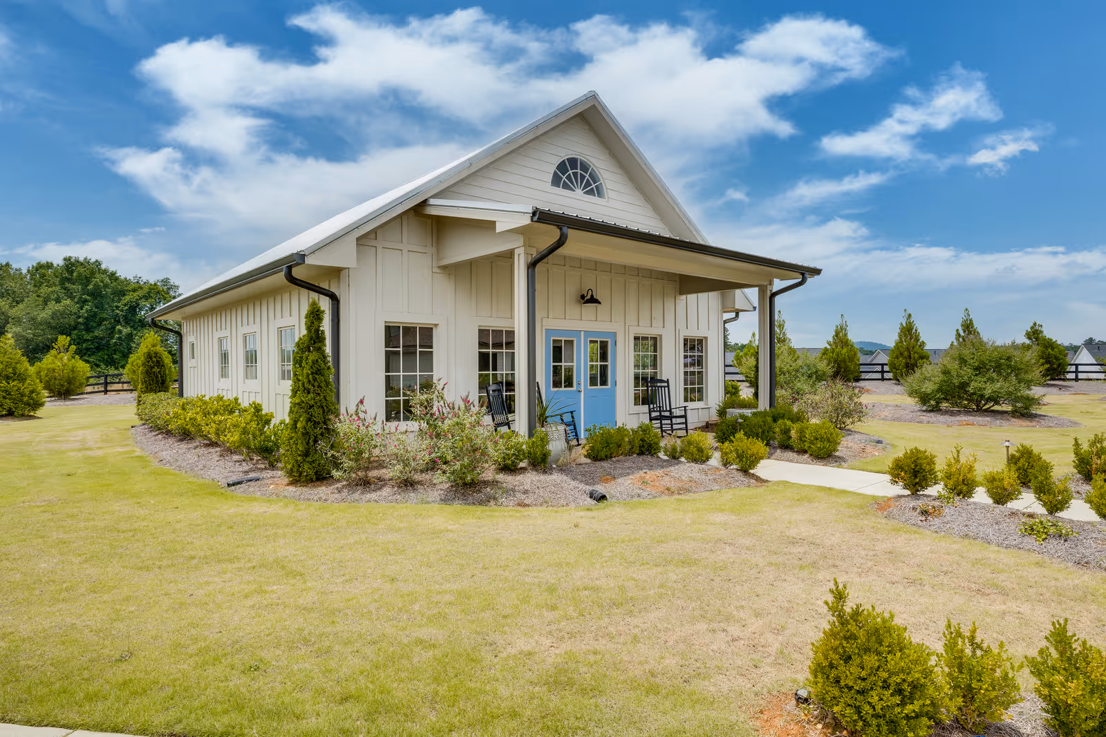 A single-story building with white siding and a blue double door entrance, surrounded by neatly trimmed bushes and green grass under a partly cloudy blue sky. There are two black rocking chairs on the small porch at the entrance.