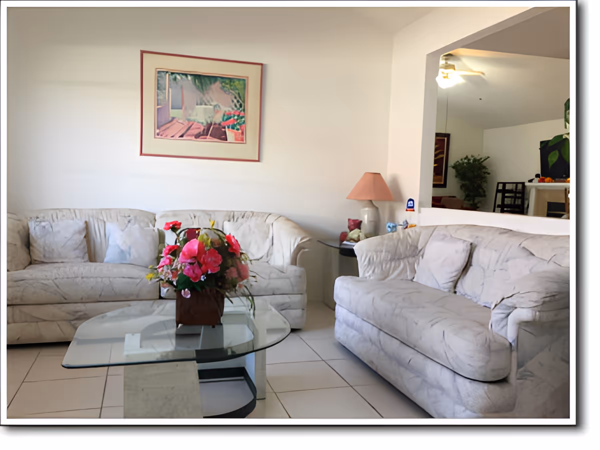 A living room with two light-colored sofas facing each other, a glass coffee table with a flower arrangement in the center, a side table with a lamp, and a framed painting on the wall. The room has tiled flooring and a partial view into another room with a ceiling fan and additional furniture.