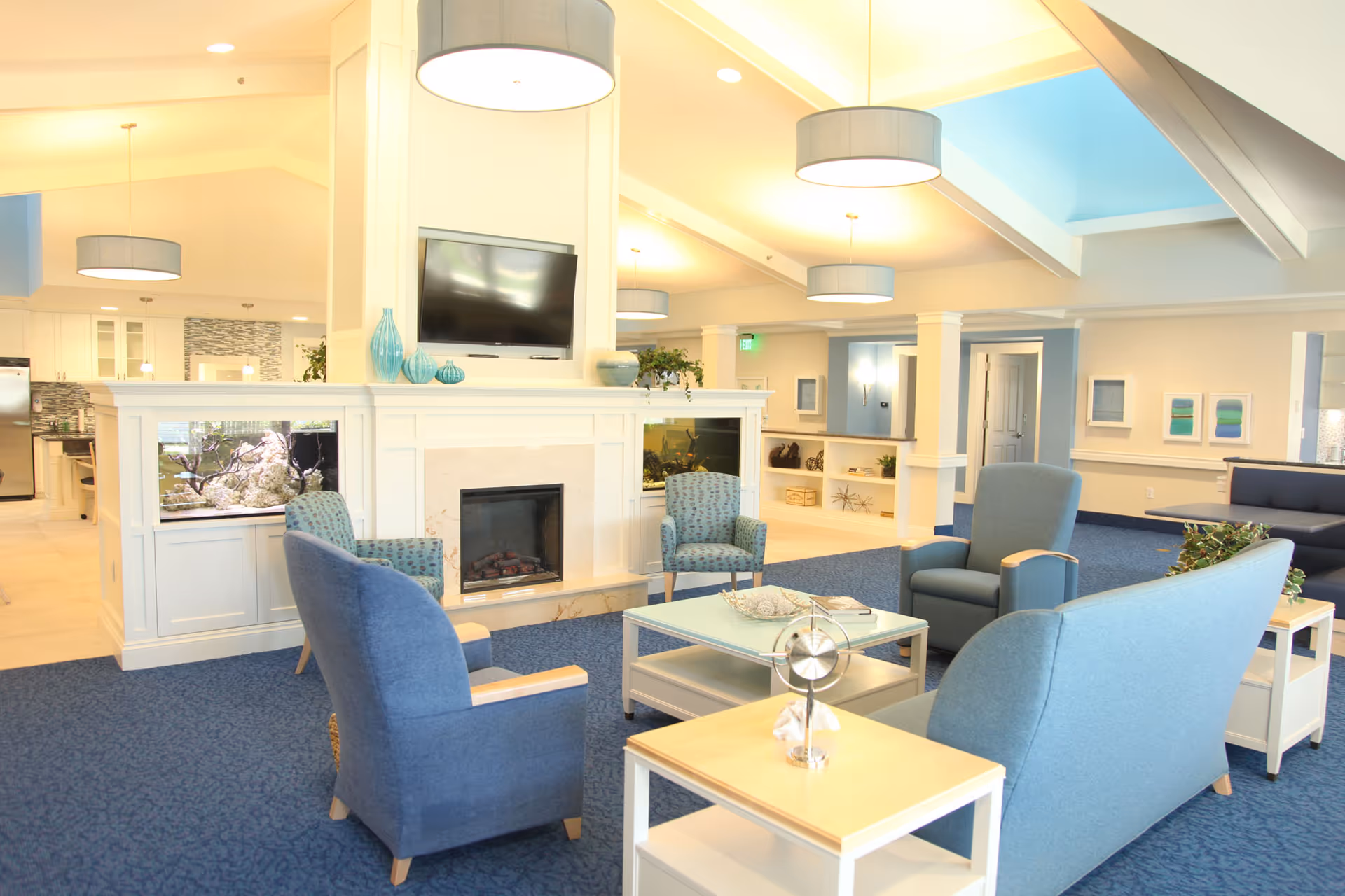 A bright and spacious living room area with blue carpet and comfortable blue upholstered chairs and sofas arranged around a glass coffee table. The room features a white fireplace with a mounted flat-screen TV above it, flanked by built-in aquariums. Modern pendant lights hang from the vaulted ceiling, and the walls are decorated with framed artwork. The background shows an open kitchen area and hallway.