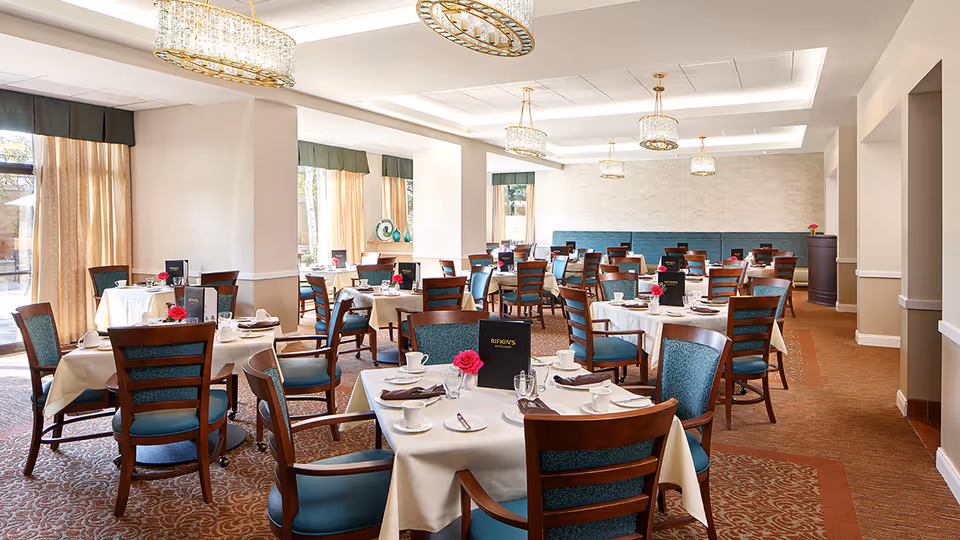 A bright and elegant dining room with multiple round tables covered in white tablecloths, each set with cups, plates, napkins, and menus. The chairs have wooden frames with teal upholstered seats and backs. Large windows with beige curtains allow natural light to fill the room, and crystal chandeliers hang from the ceiling.