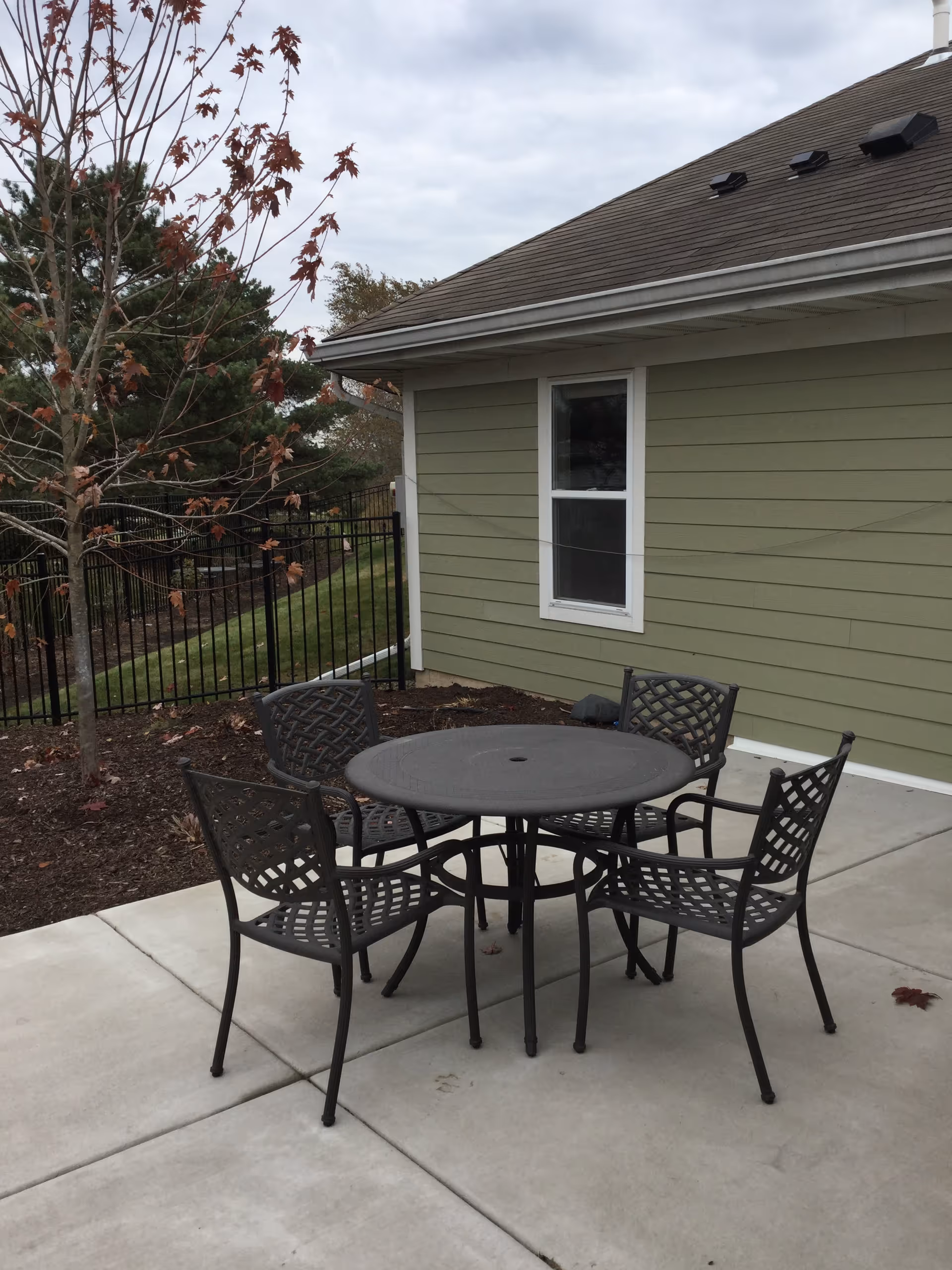 Outdoor patio area with a round metal table and four matching metal chairs on a concrete surface next to a green building with a window. There is a tree with some autumn leaves and a black metal fence in the background under a cloudy sky.