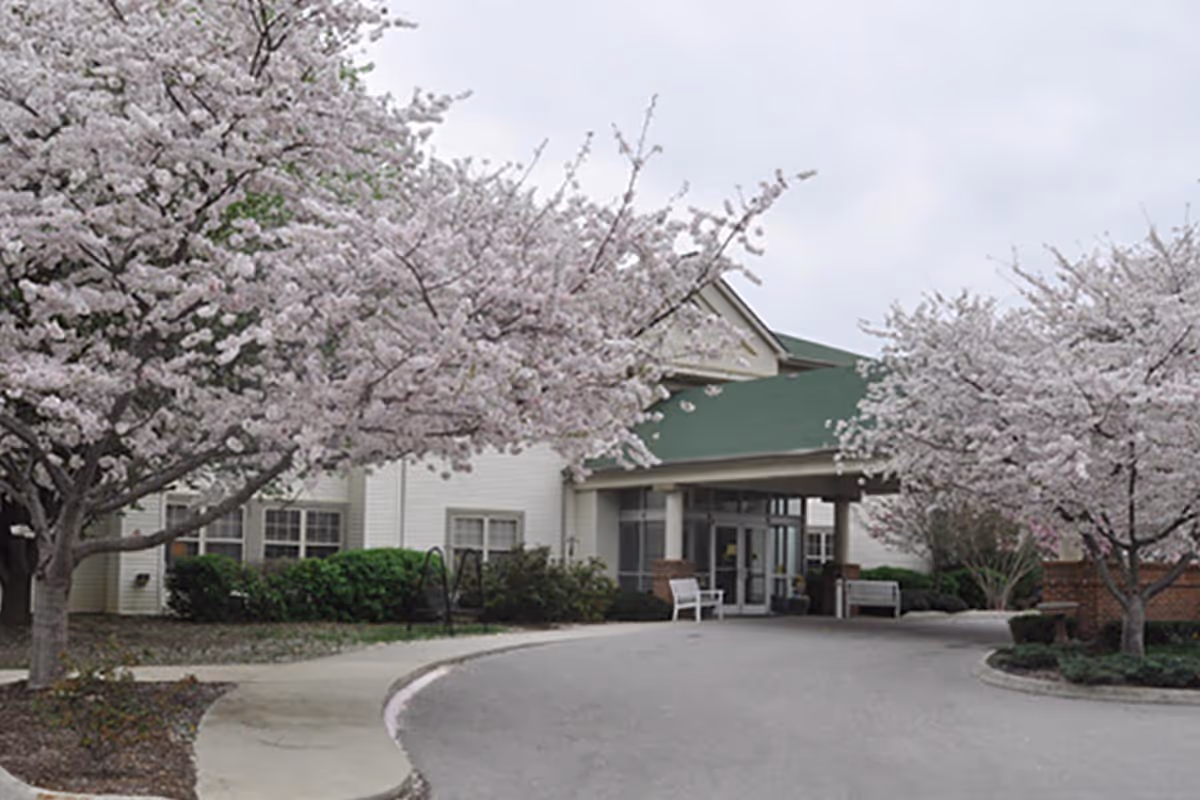 Entrance of a senior living facility with a green roof and white exterior walls, surrounded by blooming cherry blossom trees and a curved driveway leading to the front doors. Benches are placed near the entrance.