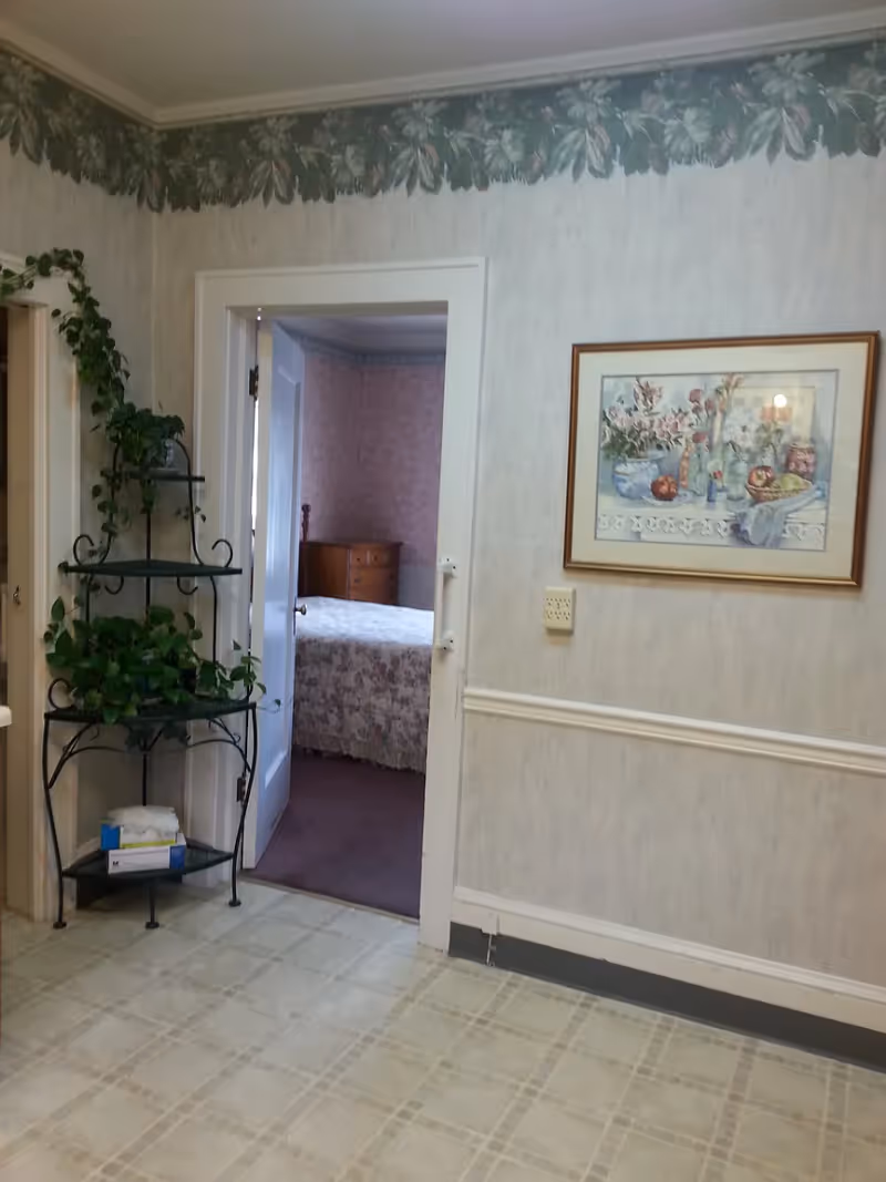 Interior view of a room with light-colored wallpaper and a floral border near the ceiling. A black metal plant stand with green leafy plants is positioned near a doorway leading to a bedroom with a bed covered in a floral bedspread and a wooden dresser. A framed floral painting hangs on the wall to the right.