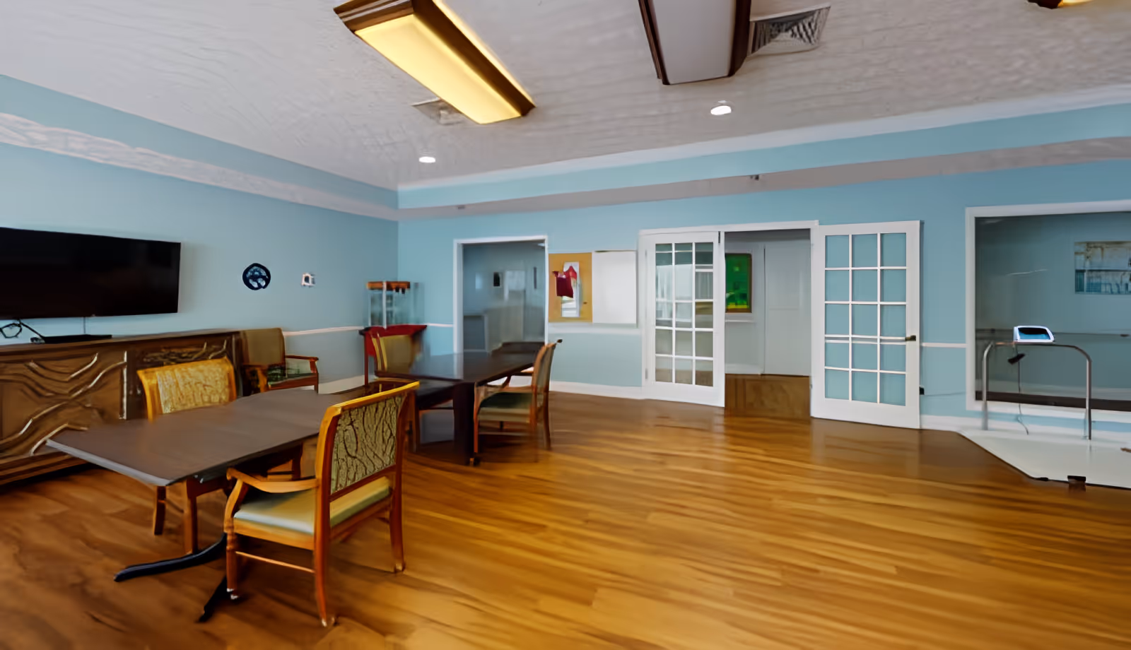 A senior living community room with tables and chairs, a wall-mounted TV, and glass double doors.