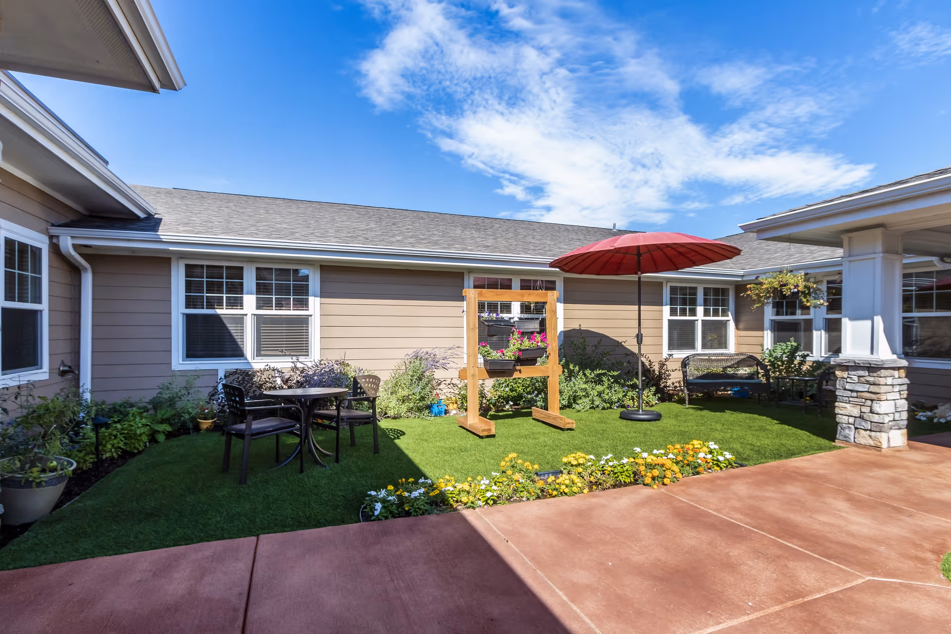 Outdoor courtyard area at Chelsea Place with a small round table and two chairs on artificial grass, a red patio umbrella, a wooden stand with flower pots, and a bench near the building. The building has beige siding and white-trimmed windows under a blue sky with some clouds.