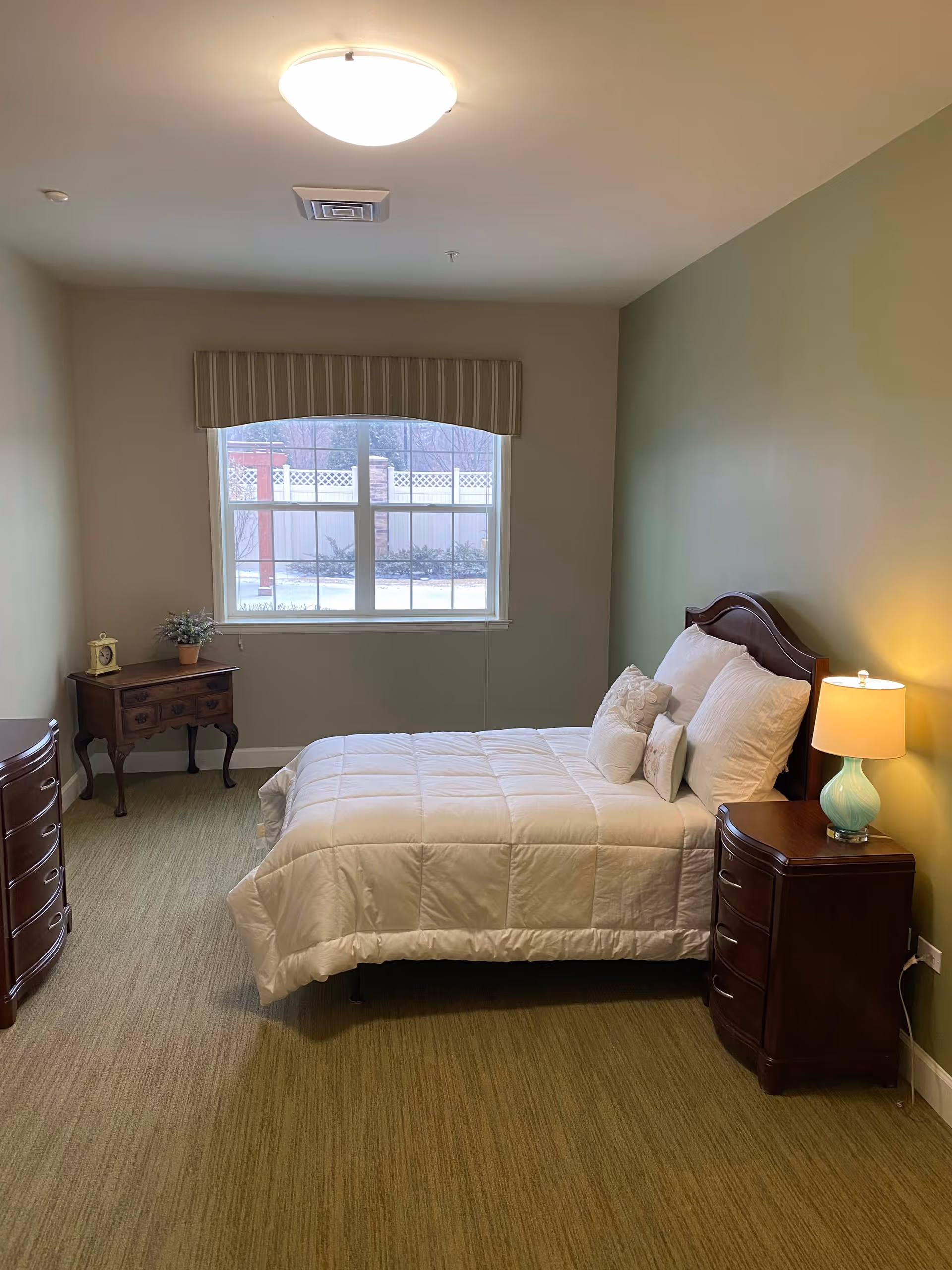 A cozy bedroom with a neatly made bed featuring white bedding and multiple pillows. The room has green carpet flooring, a green accent wall behind the bed, and a window with a striped valance letting in natural light. There are two wooden nightstands on either side of the bed, one with a small lamp and the other with a small plant and a decorative clock.