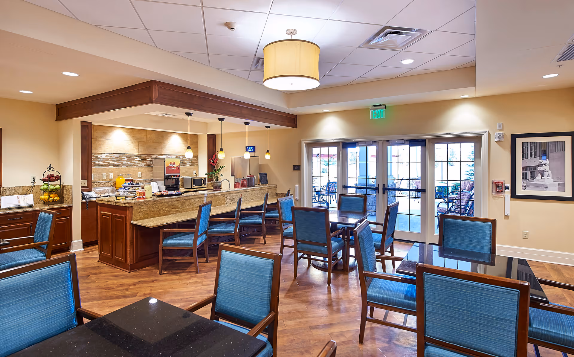 A bright and spacious dining area in a senior living facility featuring multiple tables with blue cushioned chairs, a counter with bar stools, pendant lights, and a beverage station. Large glass doors lead to an outdoor patio with additional seating.