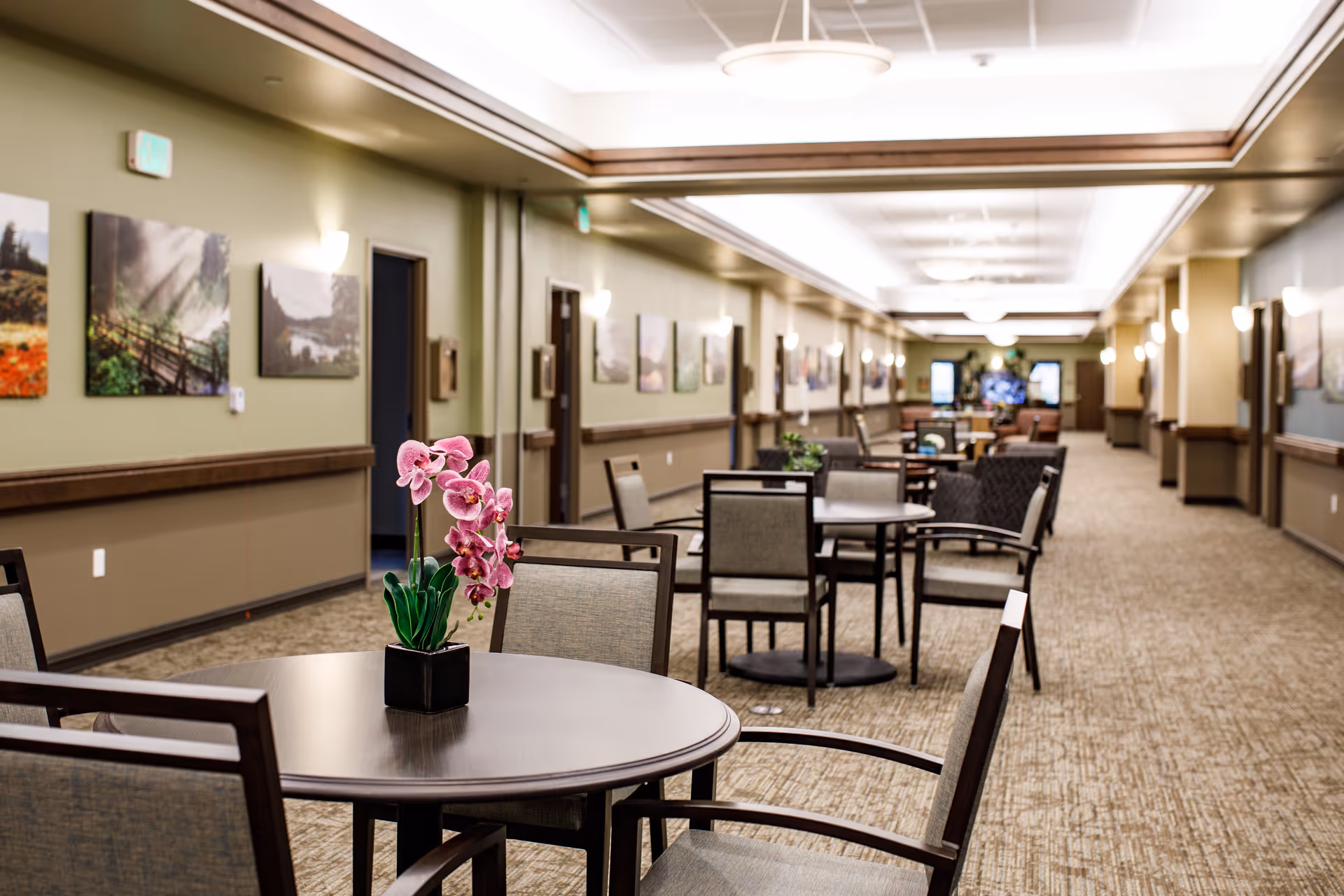 A long, well-lit hallway in a senior living facility with multiple round tables and chairs arranged along the sides. The walls are decorated with landscape paintings, and there are wall sconces providing warm lighting. A small potted orchid is placed on the nearest table.