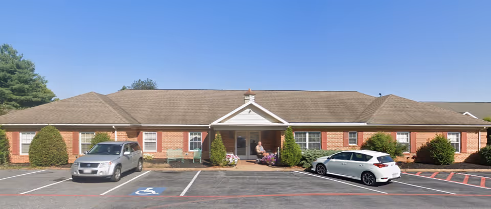 Single-story brick retirement building front with a central covered entrance, a person seated near the doorway, and cars parked in the lot with a visible handicap parking space.