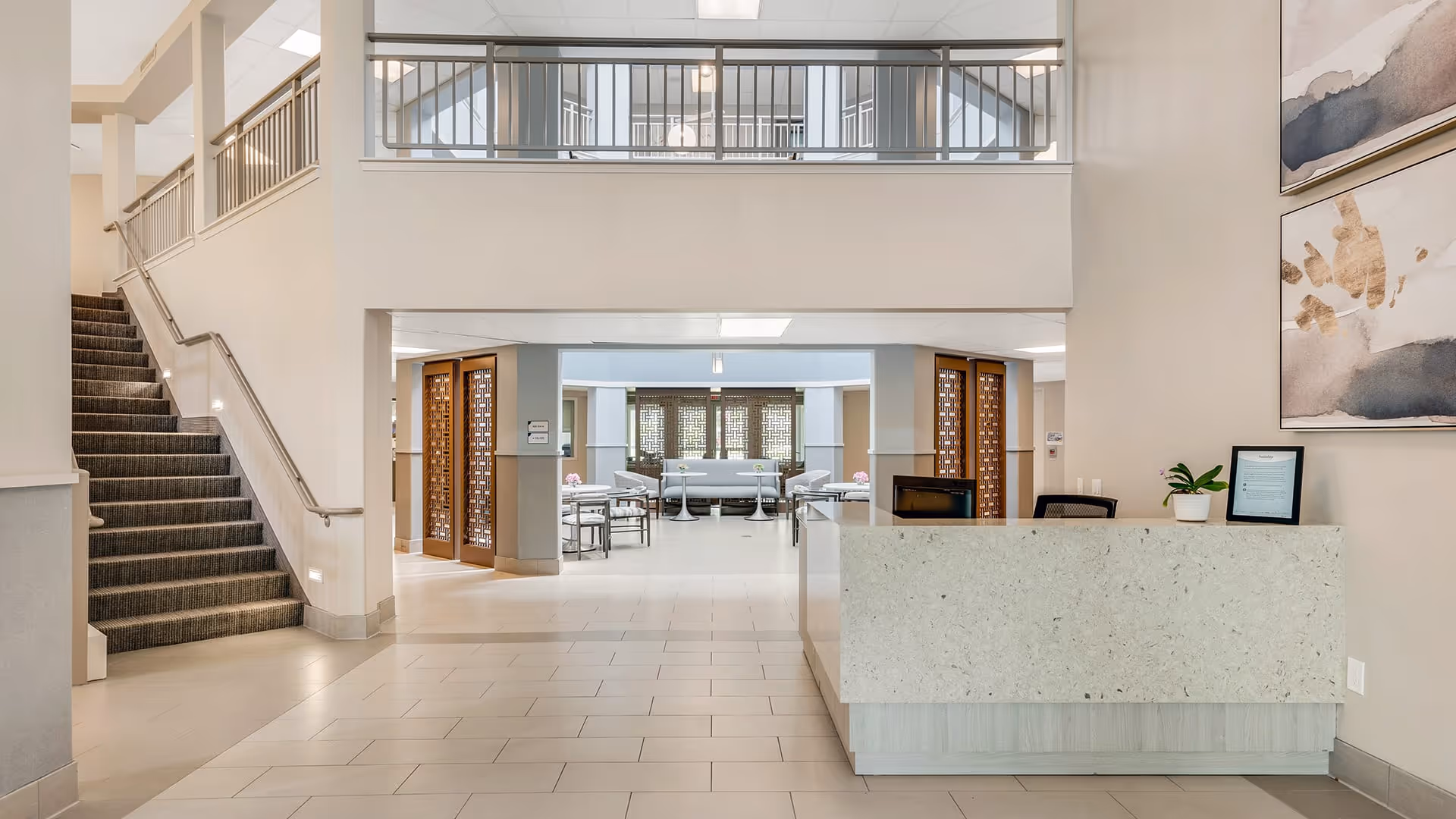 Interior view of a senior living facility lobby area with a reception desk on the right, a staircase on the left, and a seating area with tables and chairs visible in the background. The space is well-lit with neutral tones and modern decor, including abstract wall art and a small potted plant on the reception desk.
