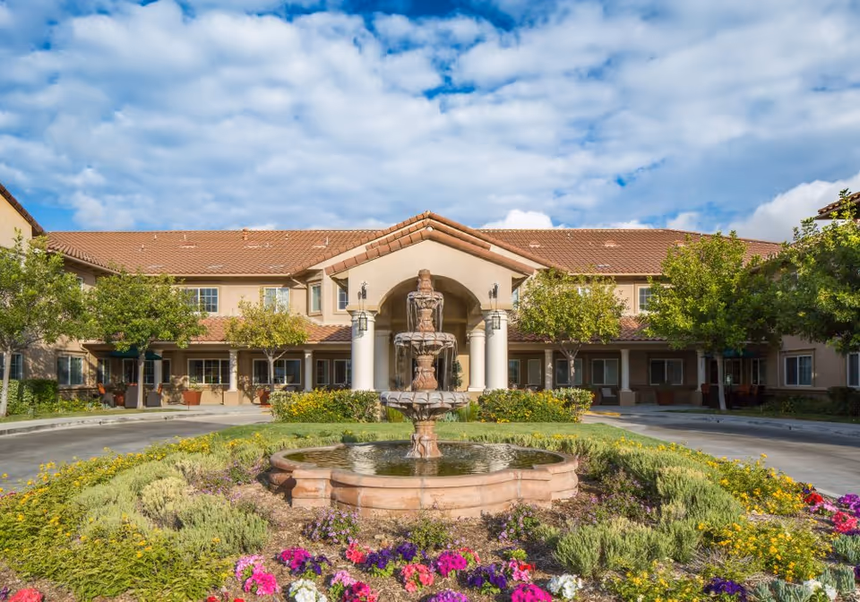 Front entrance of a two-story senior living facility with a central fountain and landscaped flowerbeds under a partly cloudy sky.