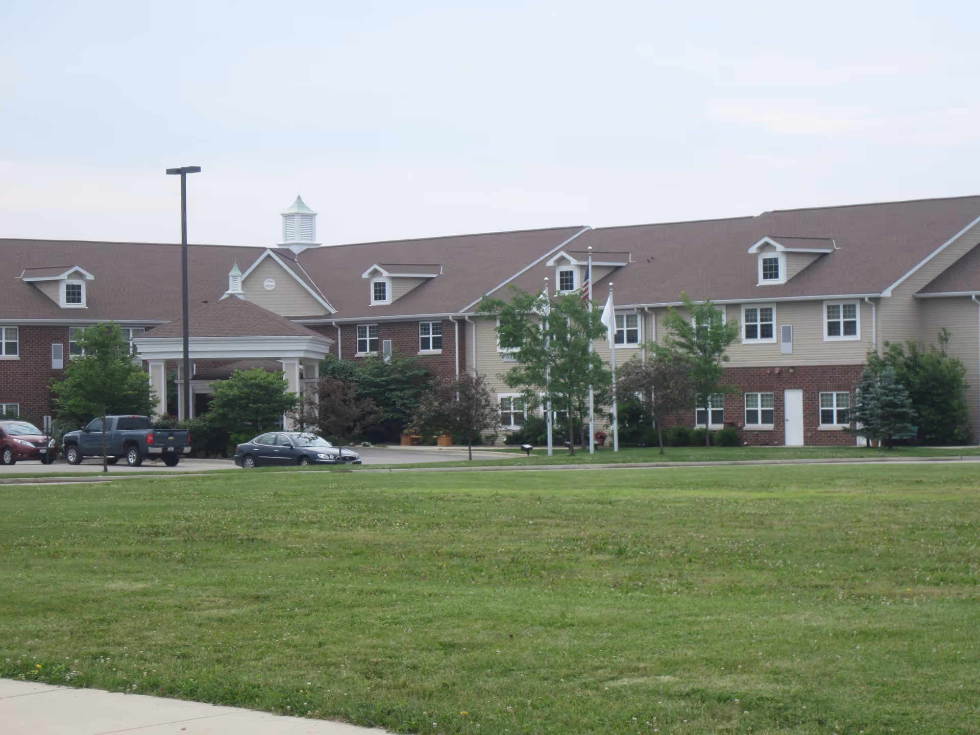 Exterior view of a large senior living facility building with beige and brick facade, multiple windows, a covered entrance, several trees, a grassy lawn in the foreground, and parked cars near the entrance.