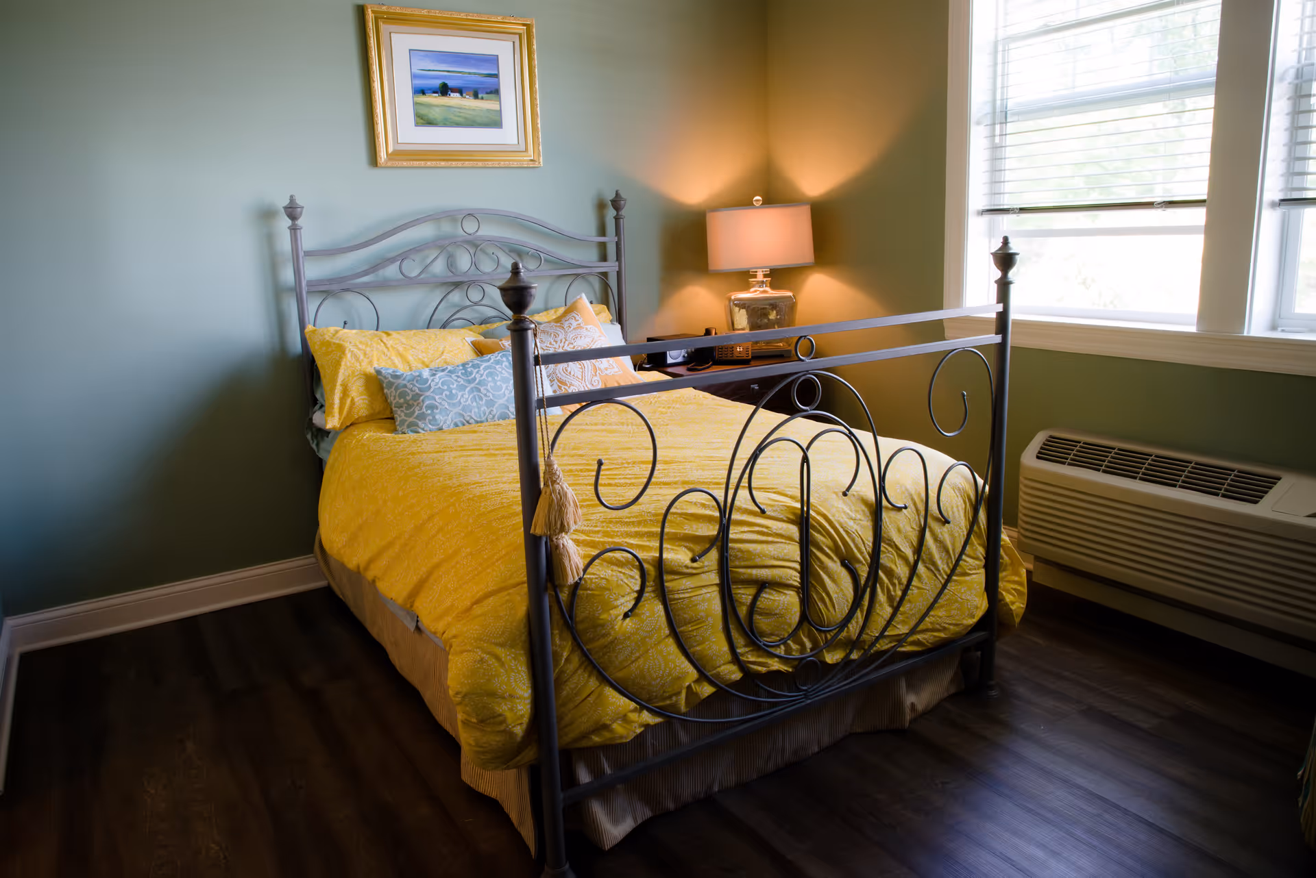 A small bedroom with an ornate metal bed dressed in yellow bedding, a bedside table and lamp, framed art on the wall, and a window with blinds.