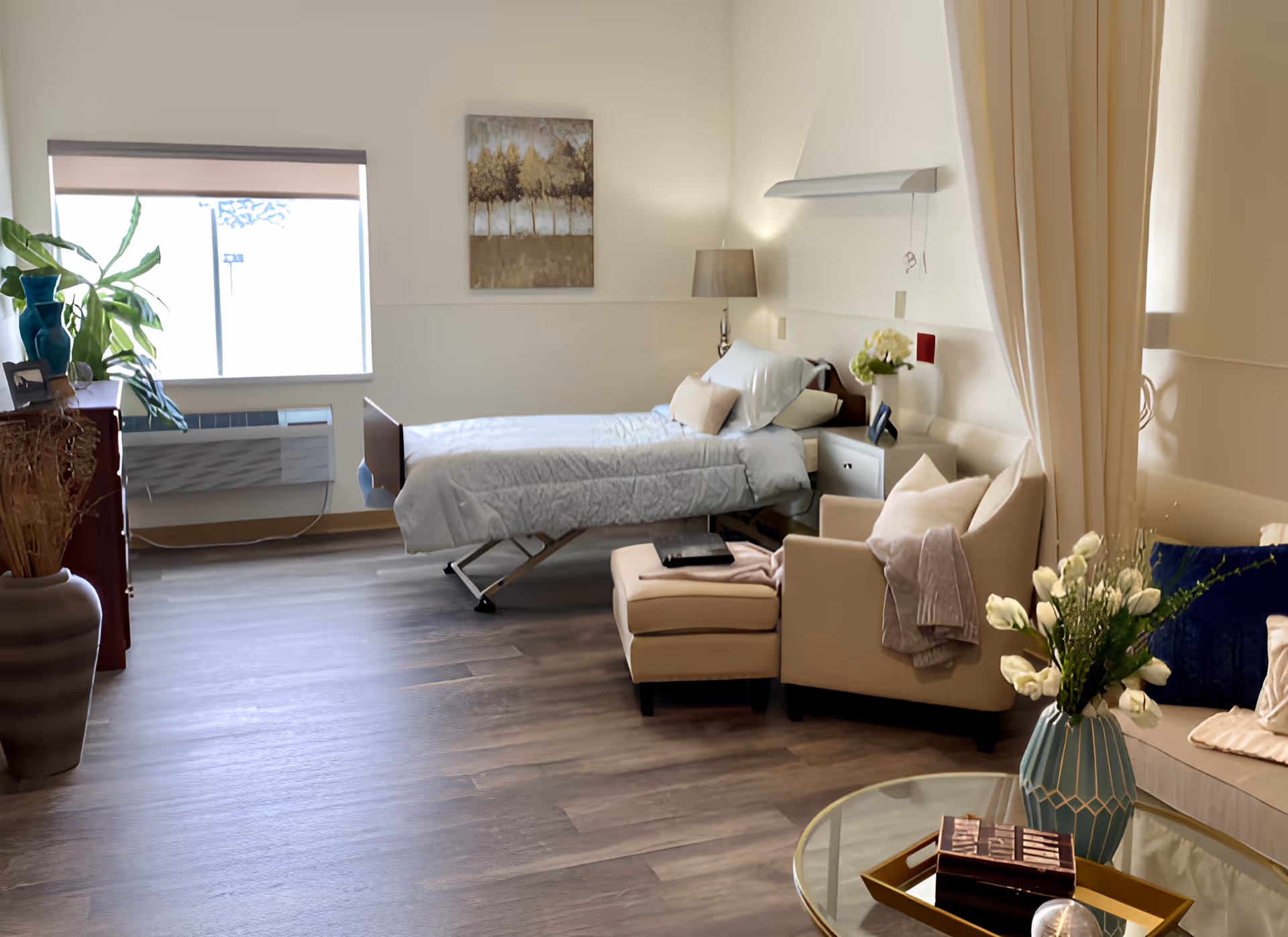 A bright and cozy room with a single hospital-style bed, a beige armchair with an ottoman, a side table with a lamp and flowers, a window with a roller shade, a dresser with plants and decor, and a glass coffee table with a vase of white flowers and a decorative box.