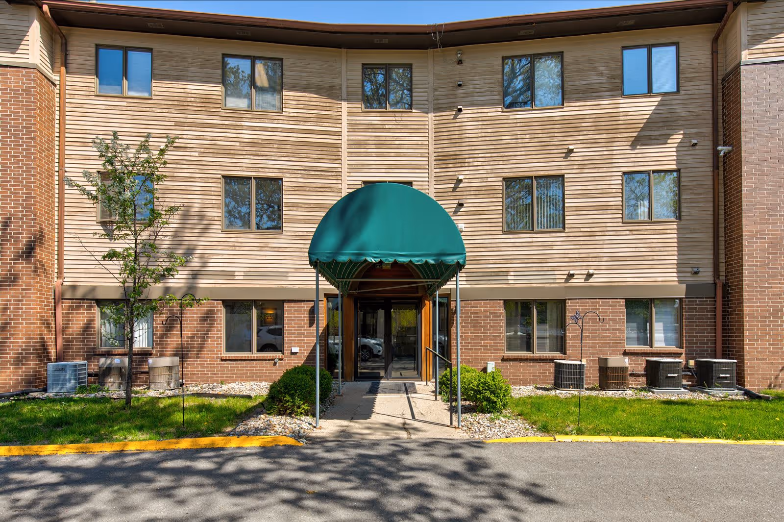 Exterior front entrance of Beaver & Adams Senior Apartments featuring a three-story building with brick and wood siding, a green canopy over the doorway, small bushes and a tree near the entrance, and several air conditioning units along the building.