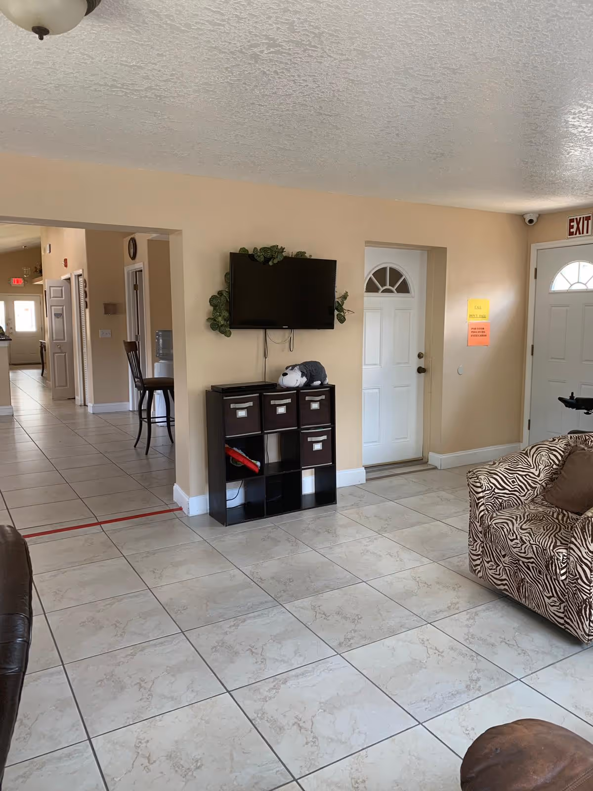 Interior view of a senior living facility common area with beige walls and tiled floor. A flat-screen TV is mounted on the wall above a black cubby storage unit with fabric bins and a stuffed animal on top. To the right, there is a white door with a window and an exit sign above it. A zebra-patterned armchair with a brown cushion is partially visible on the right side. In the background, there is a hallway with chairs and another door.