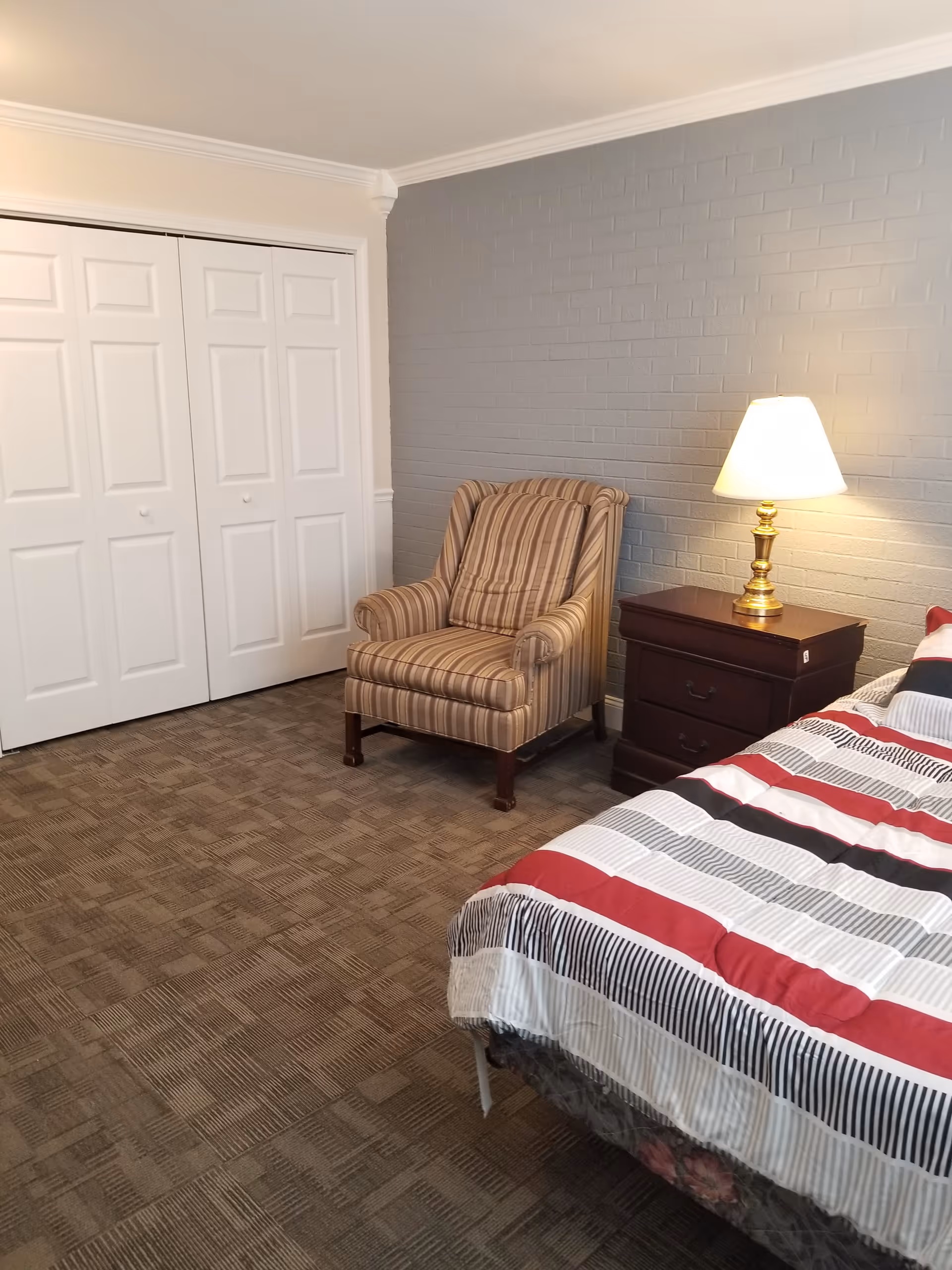 A bedroom with a striped armchair, a wooden nightstand with a brass lamp, a bed with a striped red, black, and white comforter, and white double closet doors. The walls are painted gray with a brick texture on one side.