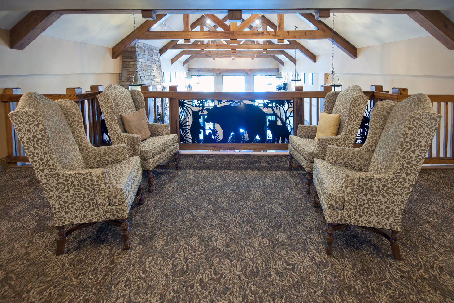 A cozy seating area with six patterned armchairs arranged in two groups facing each other on a carpeted floor. The space features wooden beams on the ceiling and a decorative railing with silhouettes of a bear and other animals. Large windows in the background allow natural light to fill the room.