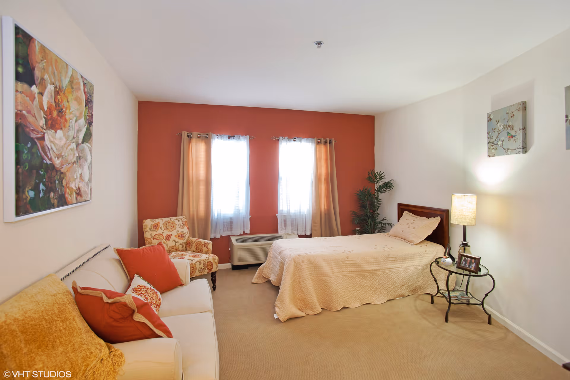 A cozy bedroom with a single bed covered in a beige quilt, a small glass side table with a lamp and photo frame, a beige sofa with orange and red cushions, a floral armchair, two windows with beige curtains, and wall art including a large flower painting and smaller framed pictures.